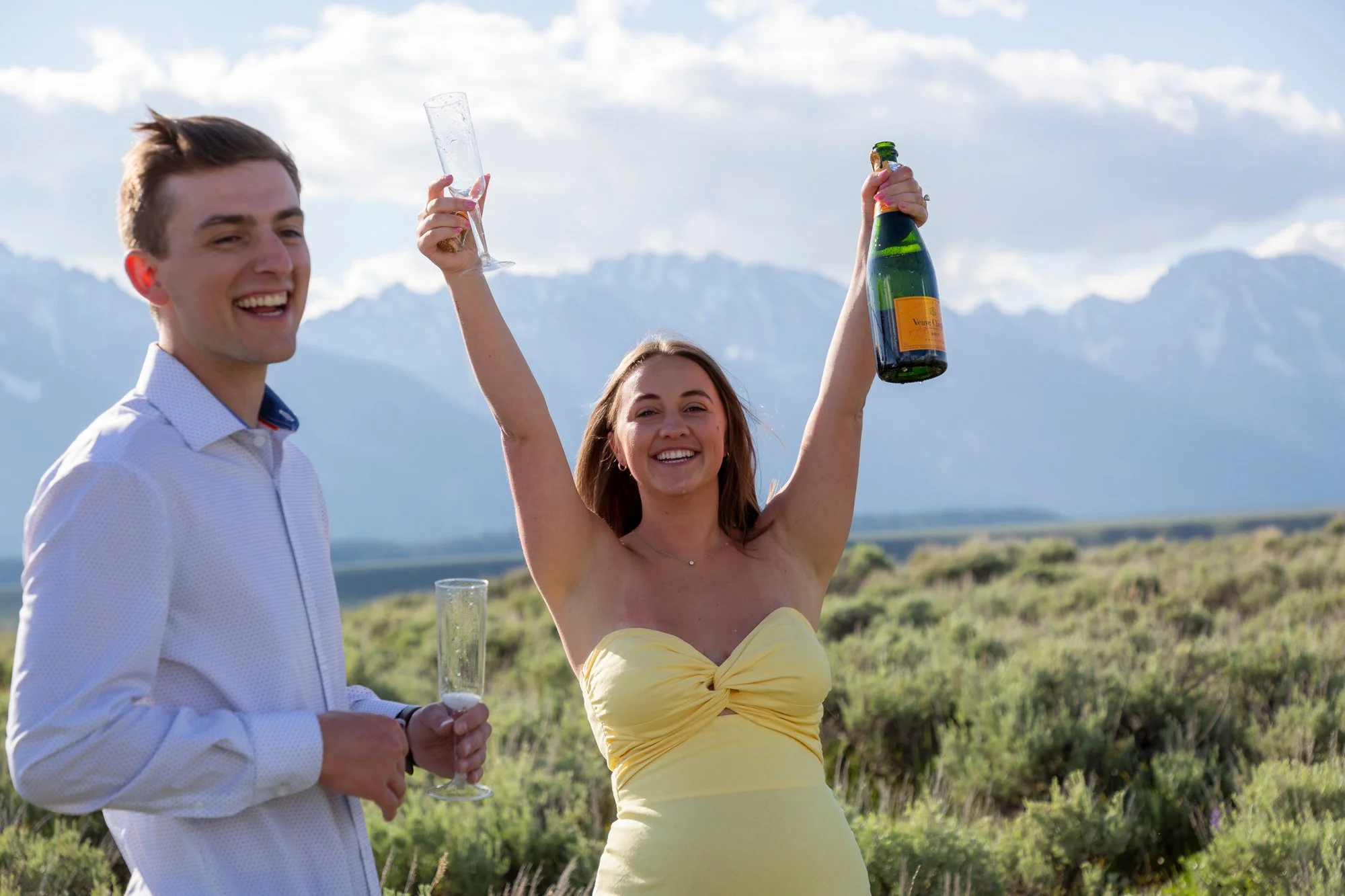 A smiling pregnant woman in a yellow dress raising a champagne bottle, and a man in a white shirt holding a champagne flute, outdoors with a mountain view in the background.