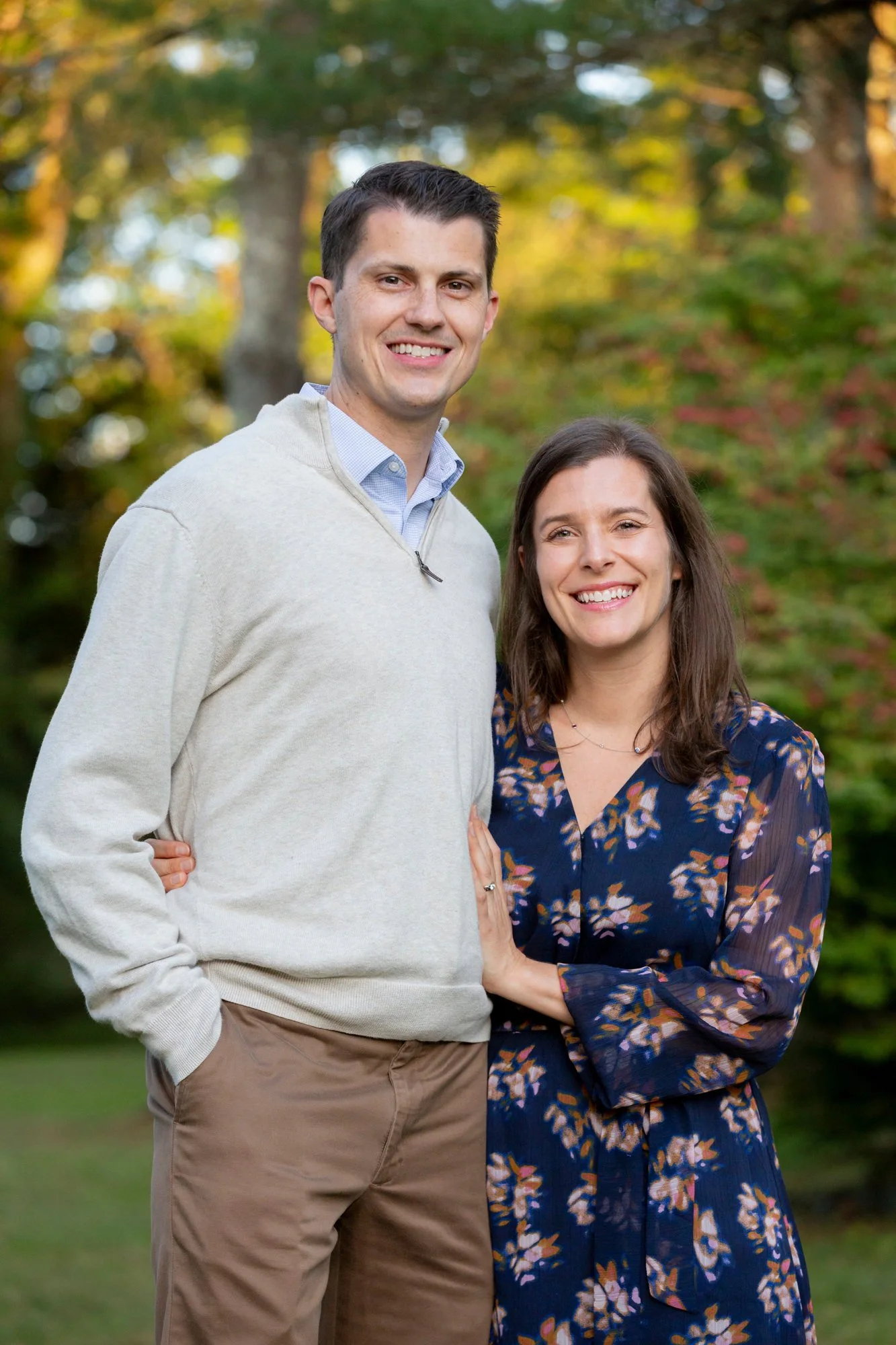 A smiling man and woman standing outdoors in front of trees with autumn foliage. The man is wearing a light-colored sweater over a collared shirt and beige pants. The woman is wearing a navy blue floral dress with long sleeves.