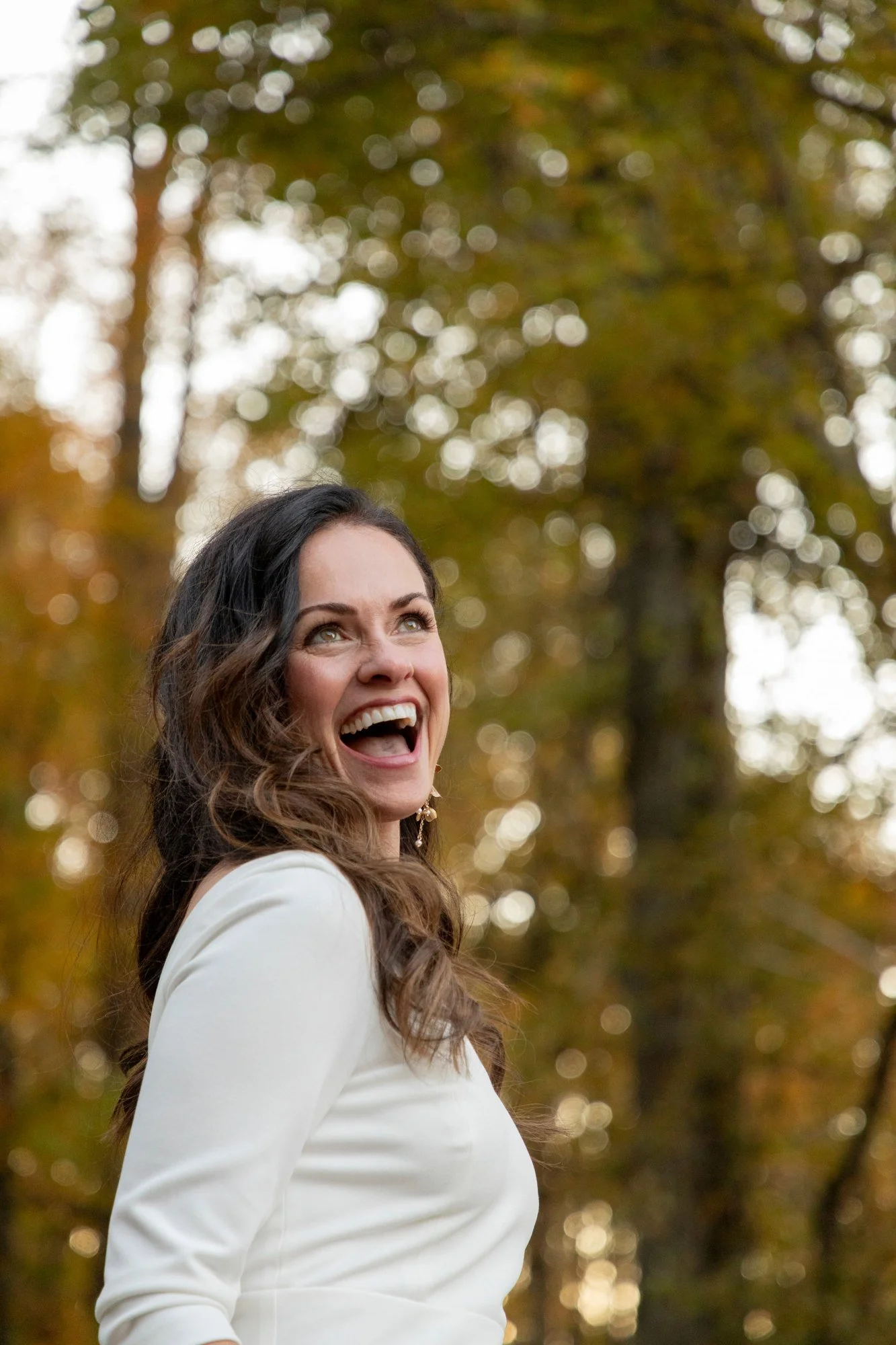 A woman with long, curly brown hair and light skin, looking up and smiling while standing outdoors with trees and sunlight in the background.