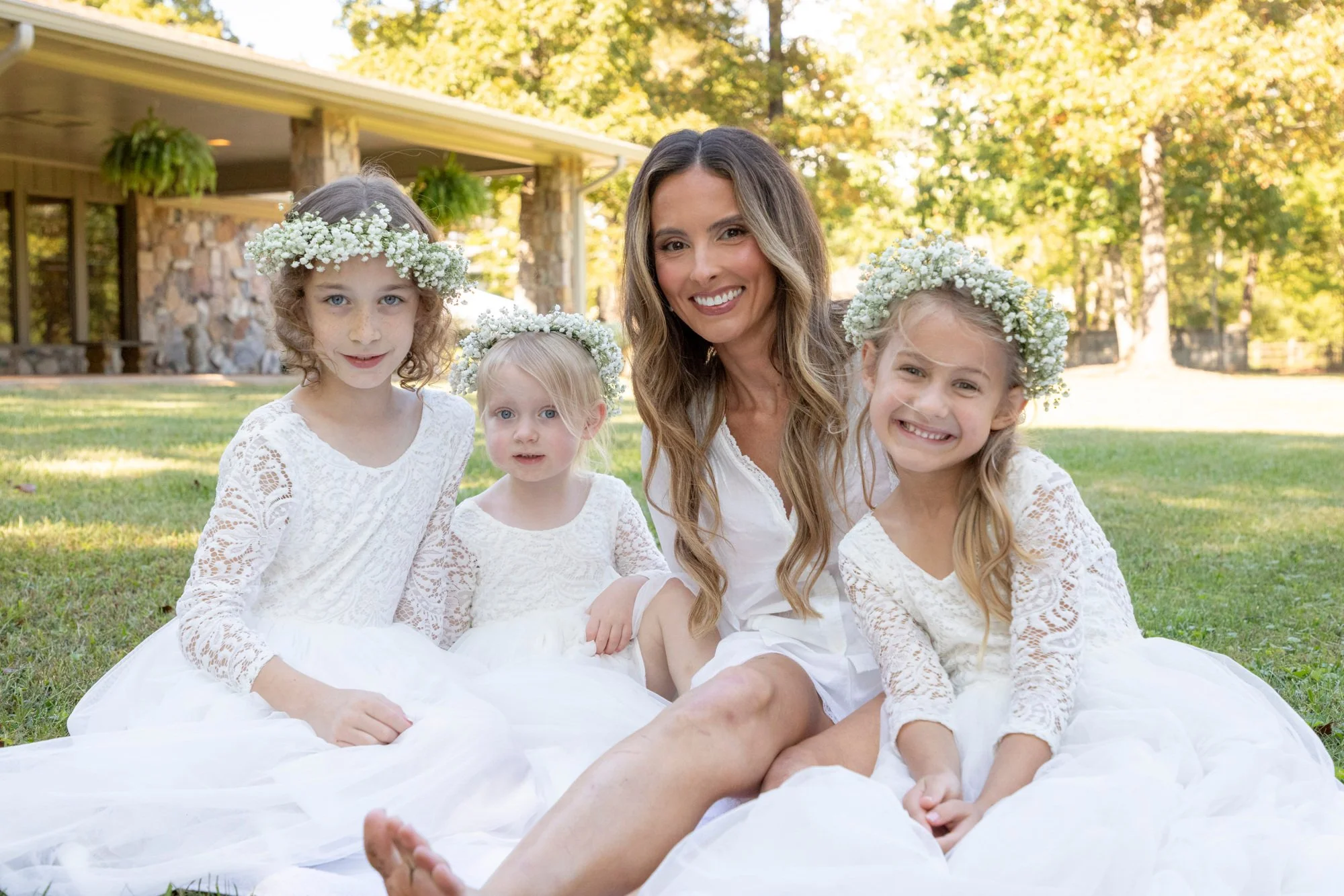 A woman and four young girls sitting on grass outside on a sunny day, dressed in white lace dresses and wearing flower crowns made of baby's breath, with a house and trees in the background.