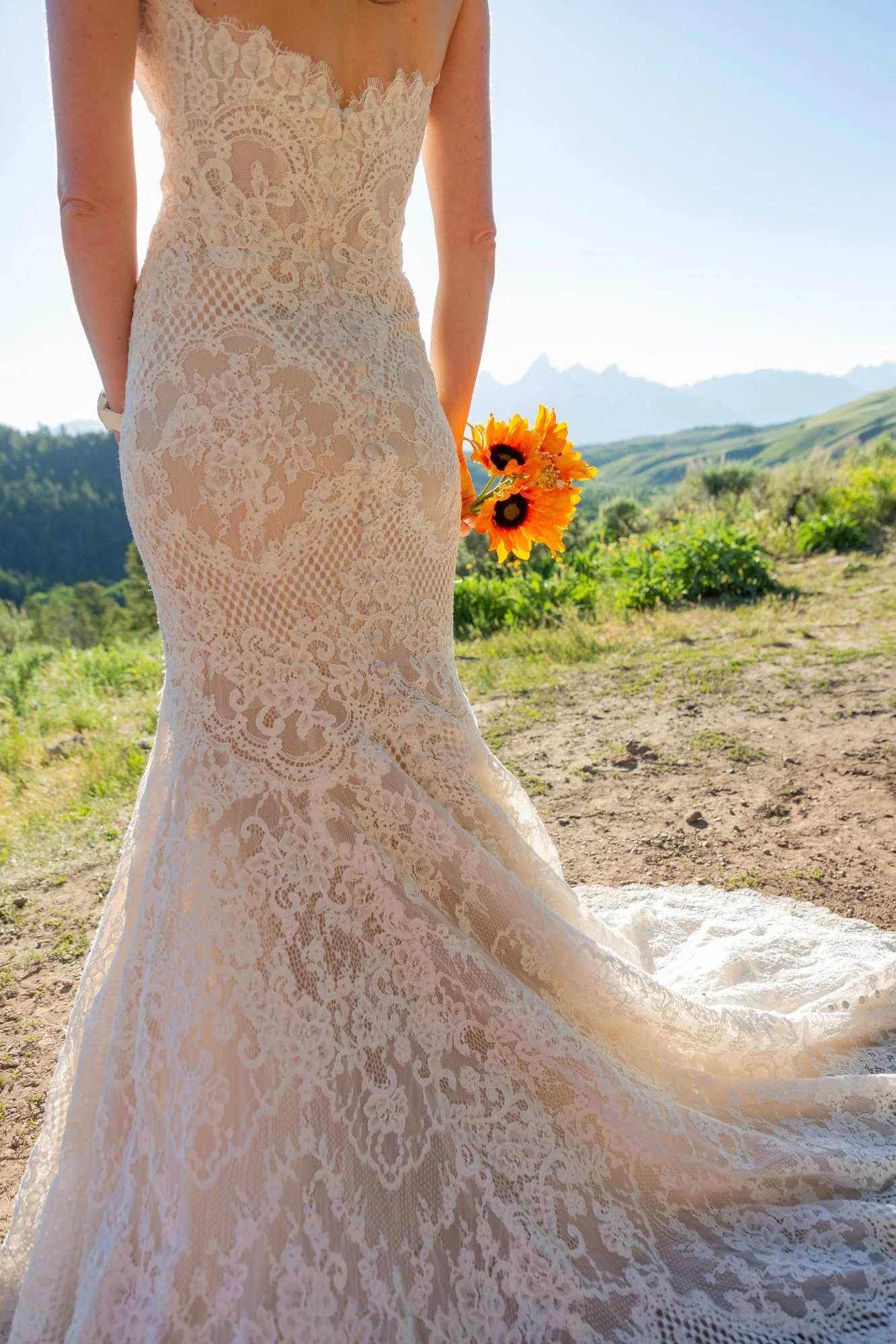 Close-up view of a person in a lace wedding dress holding a bouquet of sunflowers in an outdoor setting with mountains in the background.