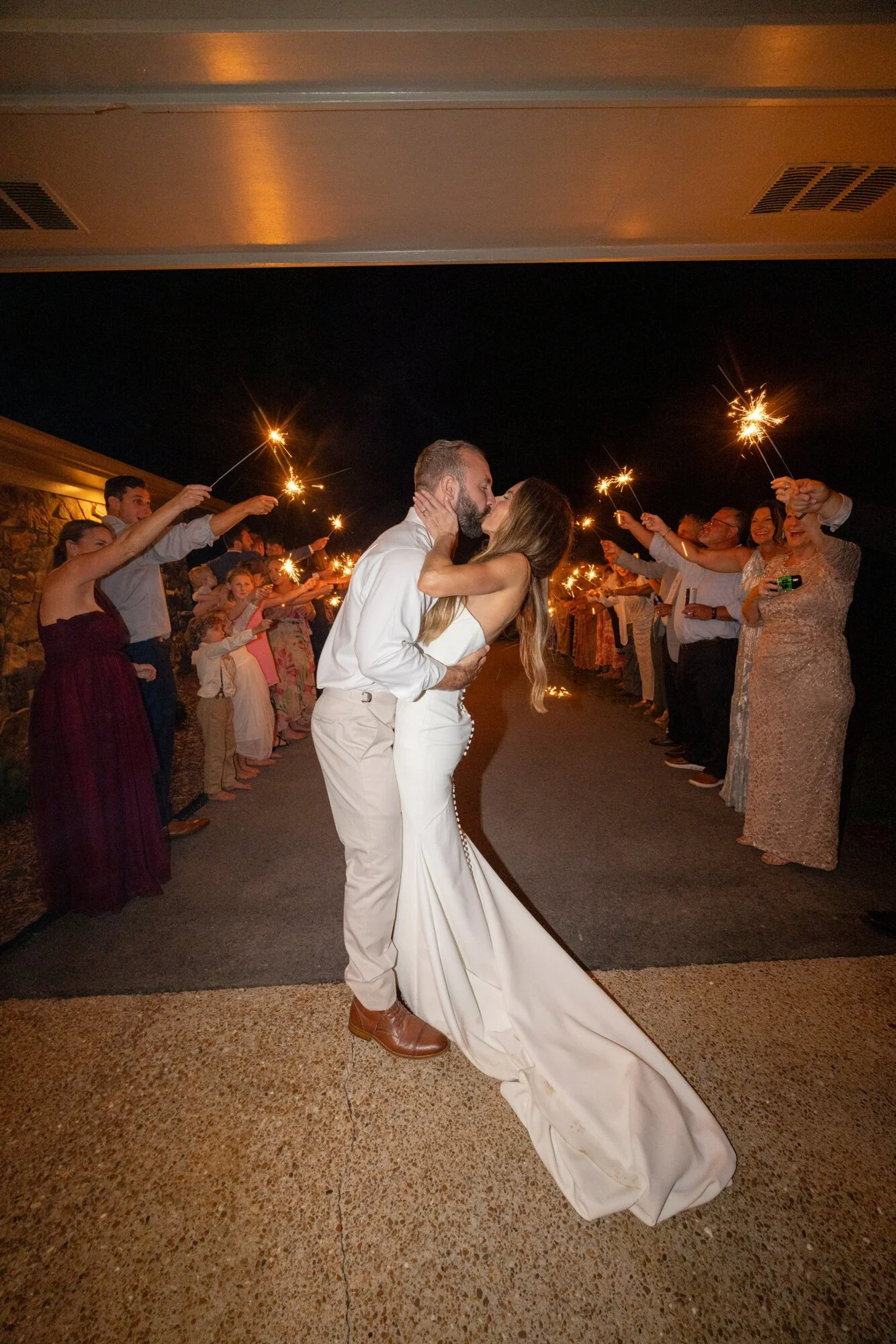 A newlywed couple shares a kiss under sparklers held by friends and family at night during a wedding celebration.