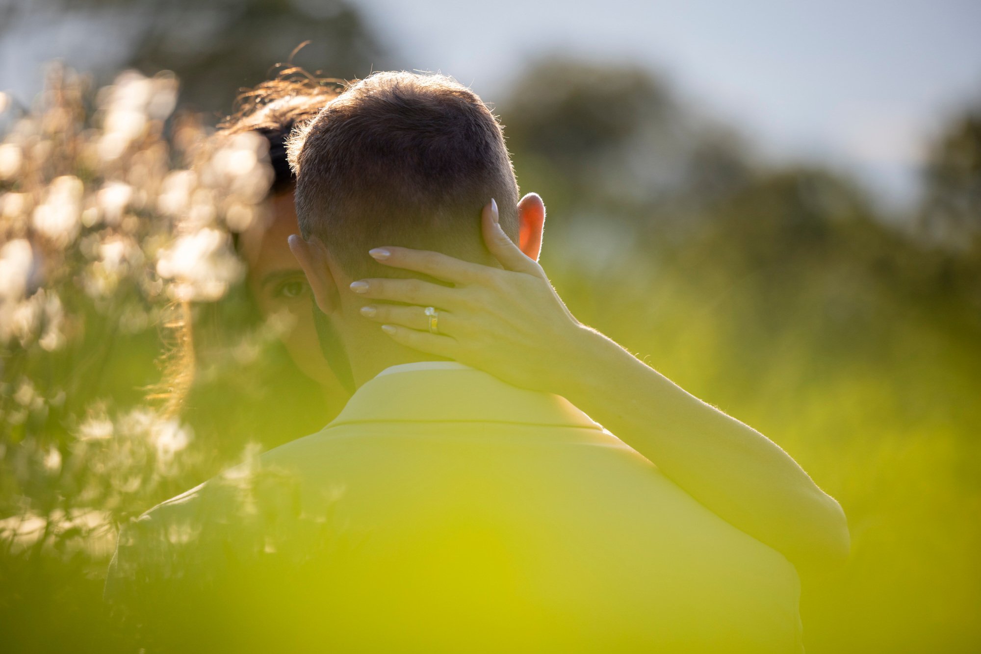 A person with short hair holding their head with both hands in an outdoor setting with greenery and sunlight.