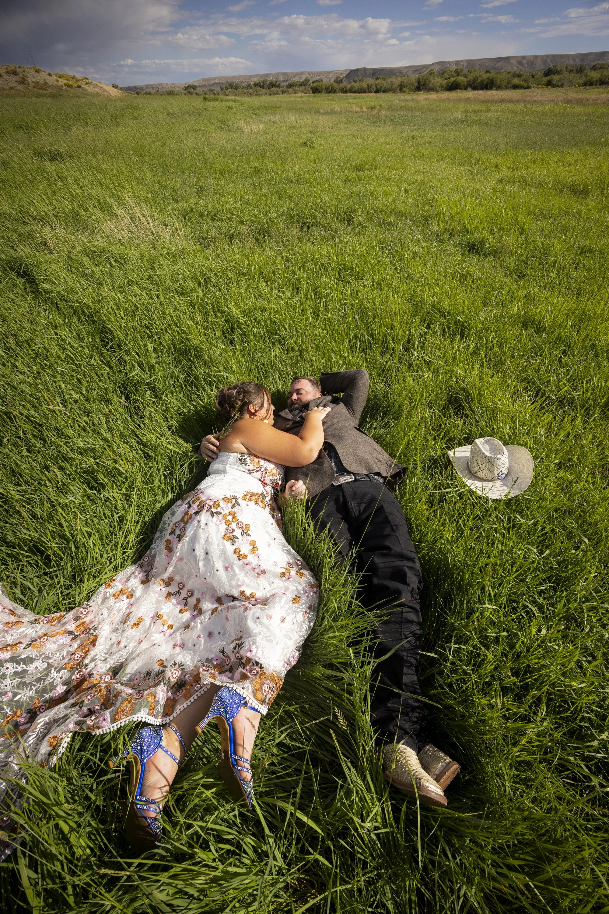 A couple lying in a grassy field, embracing each other. The woman wears a floral dress and blue high heels, and the man wears a suit and brown shoes. A sun hat is on the grass nearby. The landscape includes green grass, distant hills, and a partly cl
