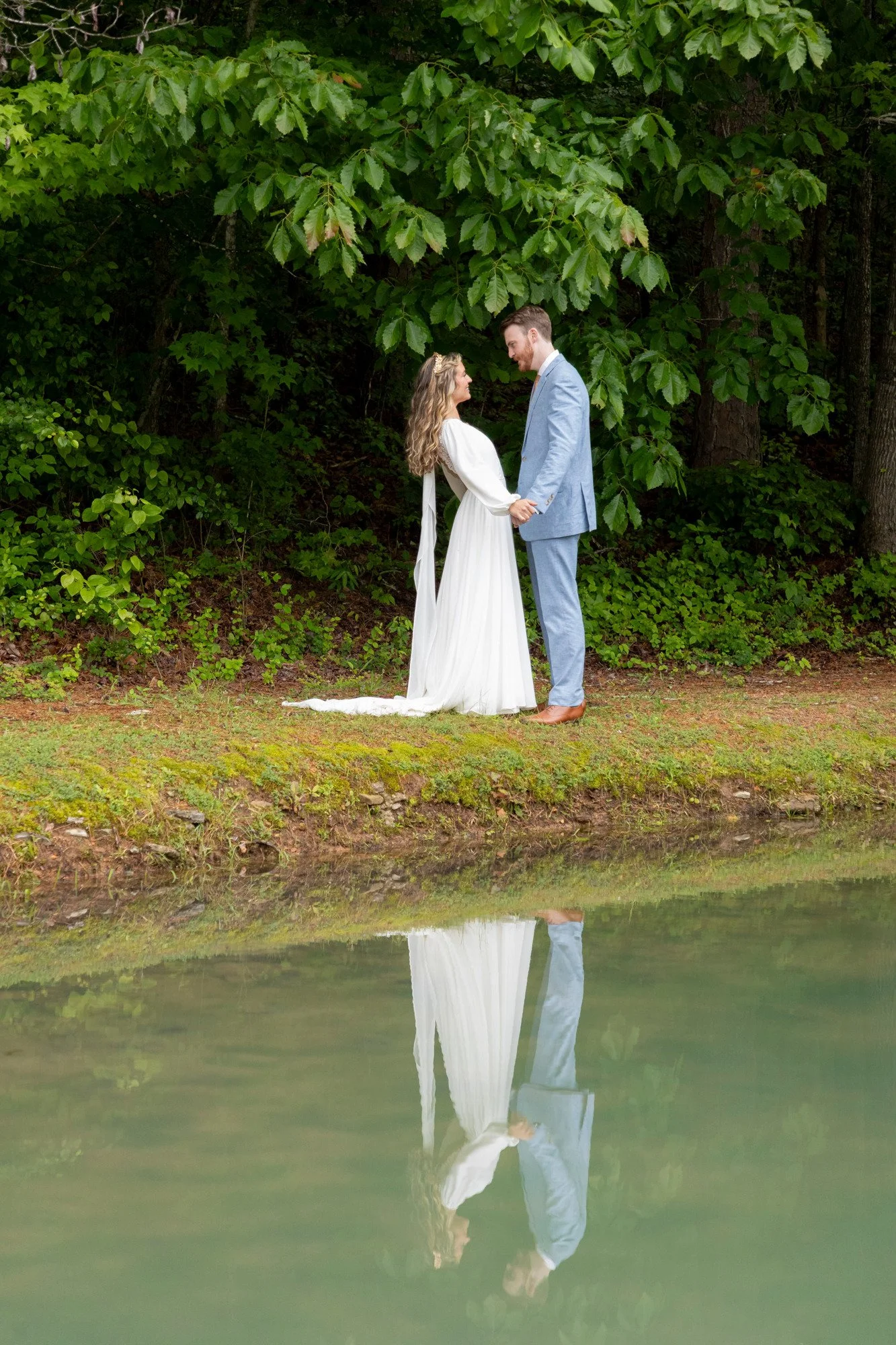 A bride and groom holding hands, standing outdoors by a lake with green trees in the background. Their reflection is visible in the water.