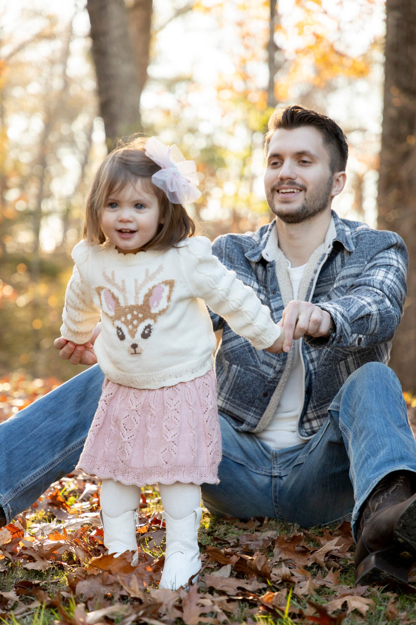 A young girl with a sweater featuring a reindeer design stands outdoors in a park with fall leaves, while a man, likely her father, sits behind her on the ground and holds her hand.