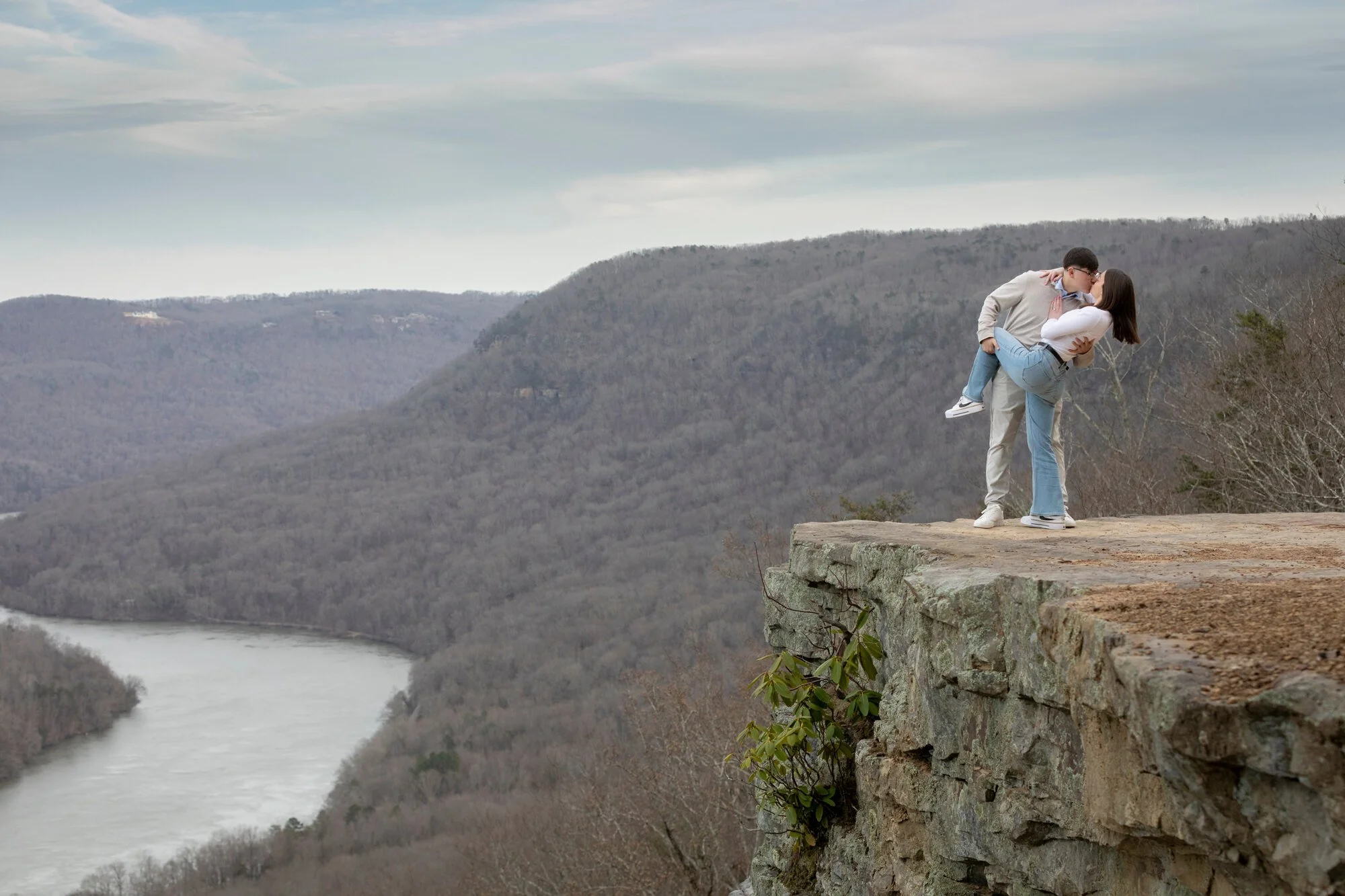 A couple standing on a rocky overlook by a river in a mountainous area, with the man lifting the woman in his arms while they kiss.