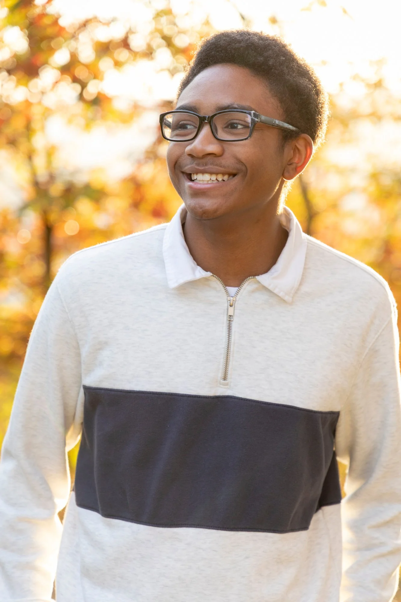 Young man with glasses, smiling outdoors in autumn, wearing a light-colored hoodie with a dark horizontal stripe, background of fall leaves.