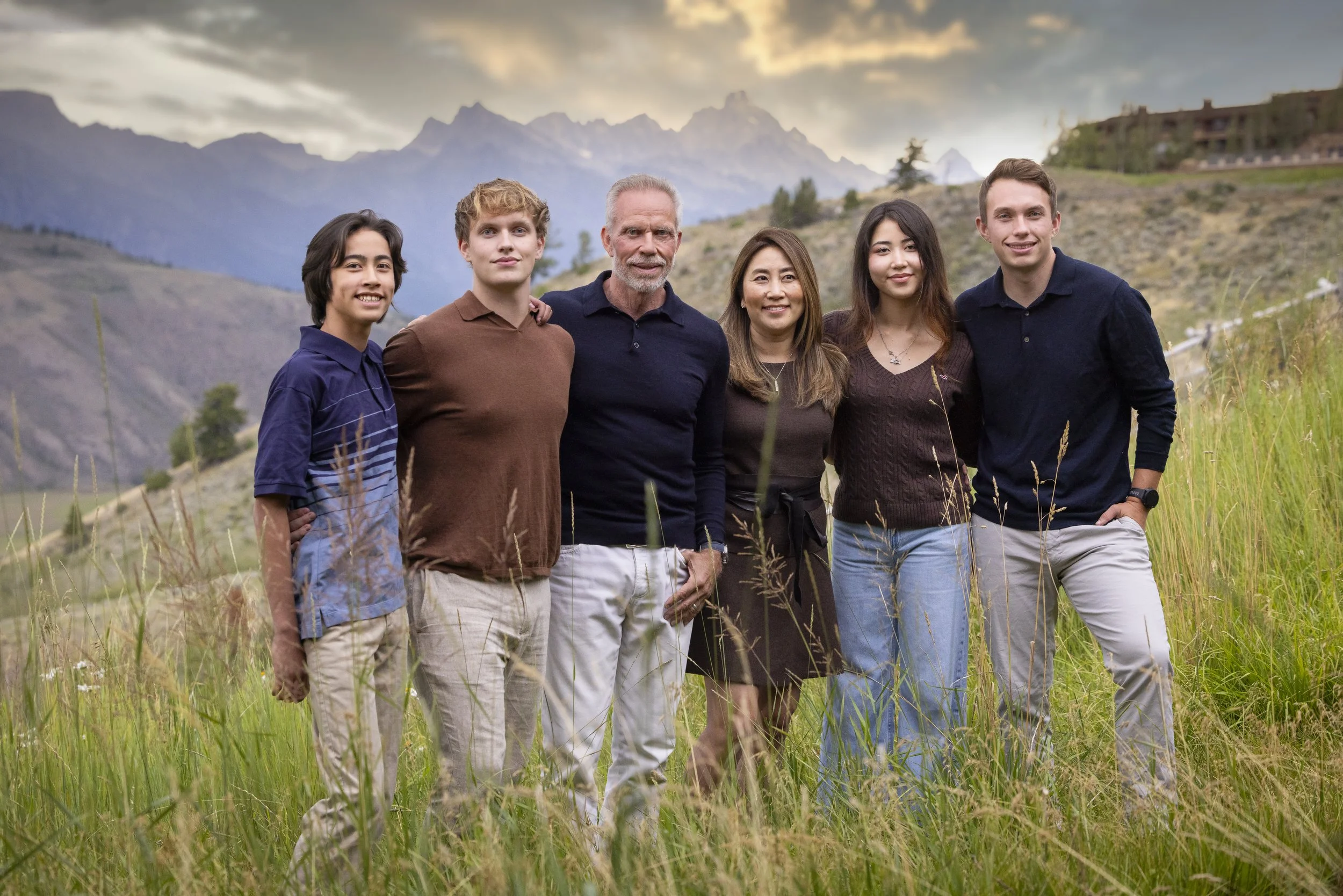 Group of six diverse people standing outdoors in a grassy field with mountains in the background, smiling at the camera.