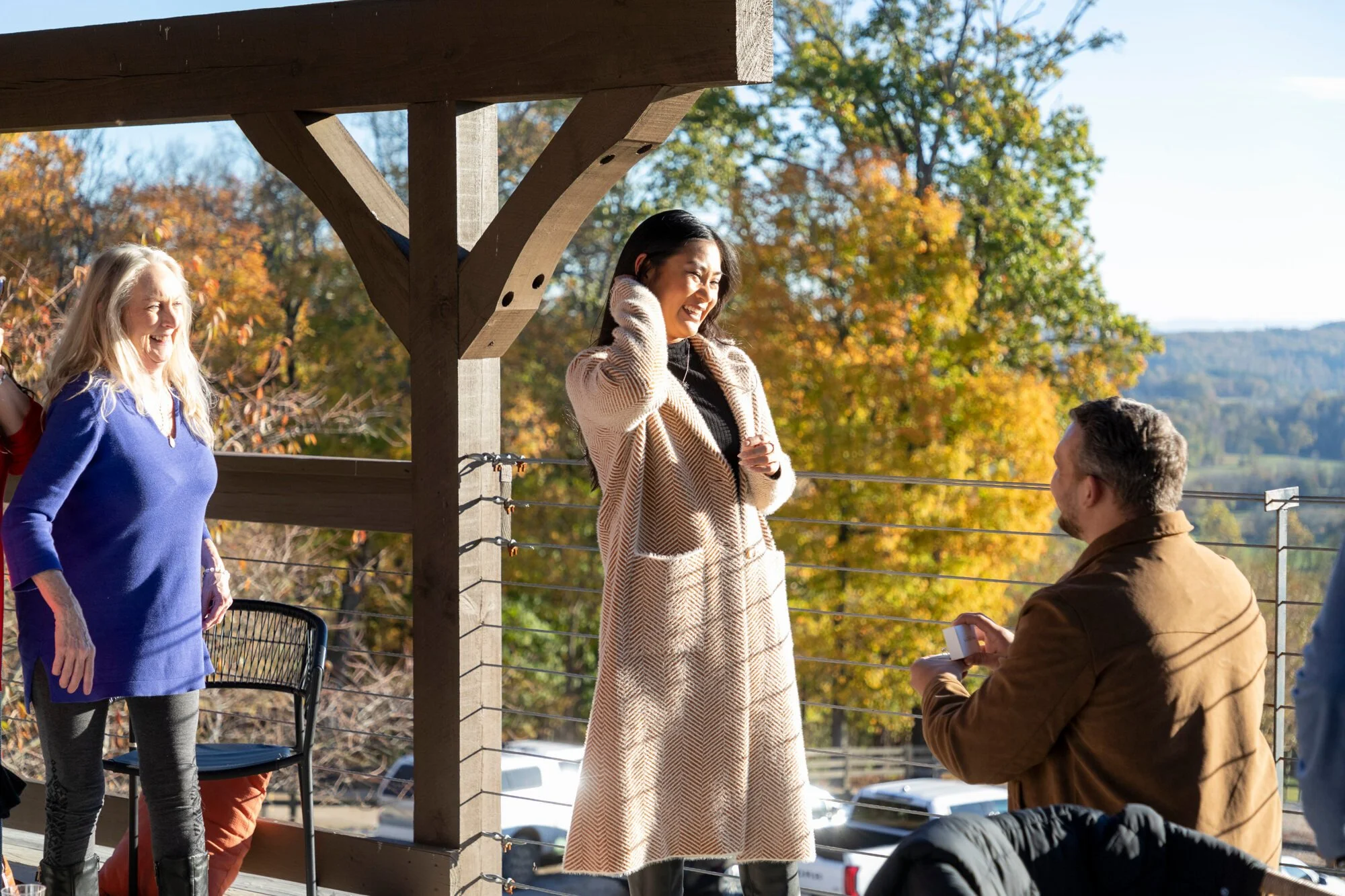 Group of people outdoors on a wooden balcony during fall, with trees showing autumn foliage in the background. A woman in a blue sweater and gray pants laughs, while a woman in a beige coat and black top smiles, talking to a man in a brown jacket who