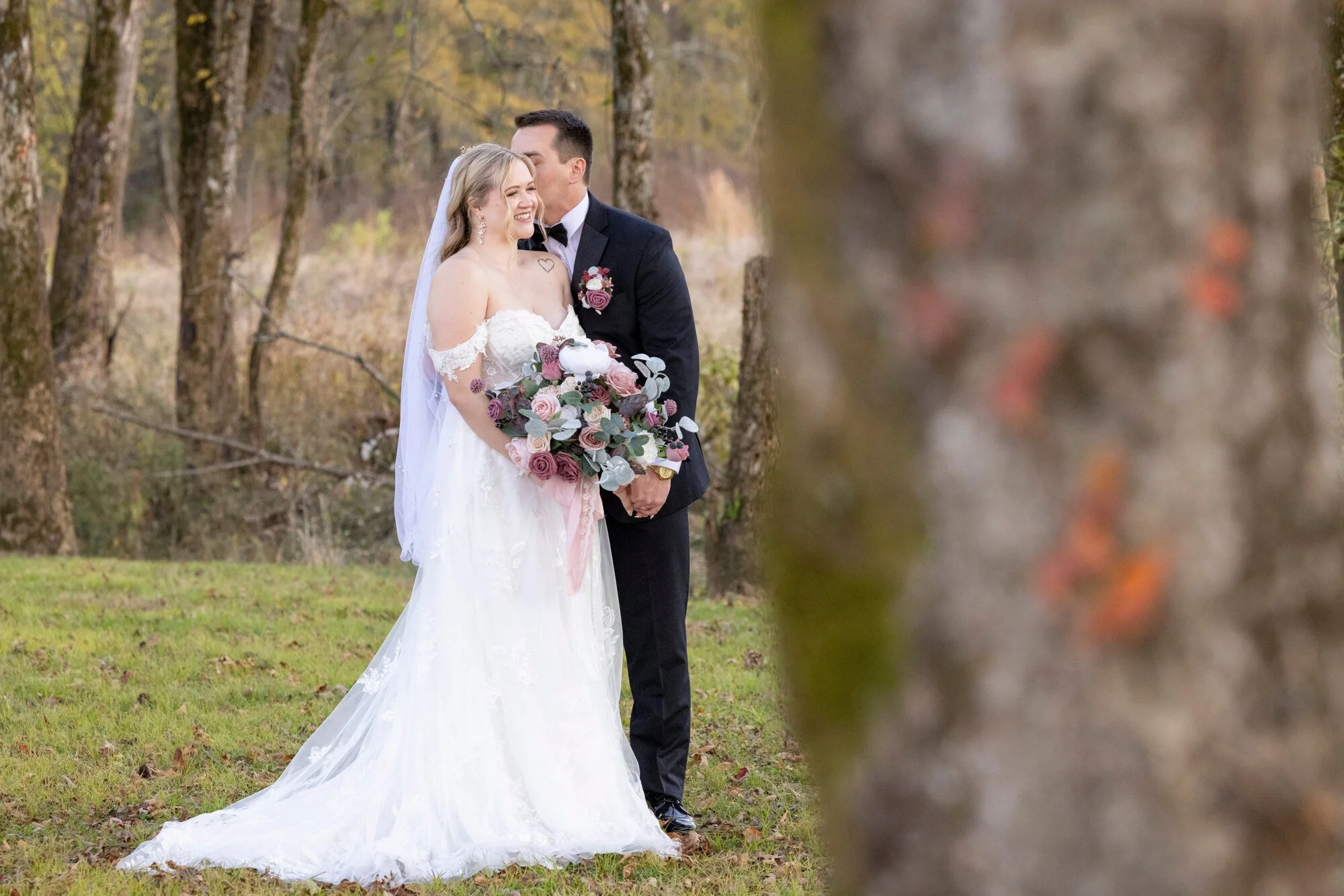 A newlywed couple stands close together in a forested area during autumn. The bride is wearing a white wedding gown with lace details and holding a bouquet of pink, purple, and green flowers. The groom is dressed in a black tuxedo with a black bow ti