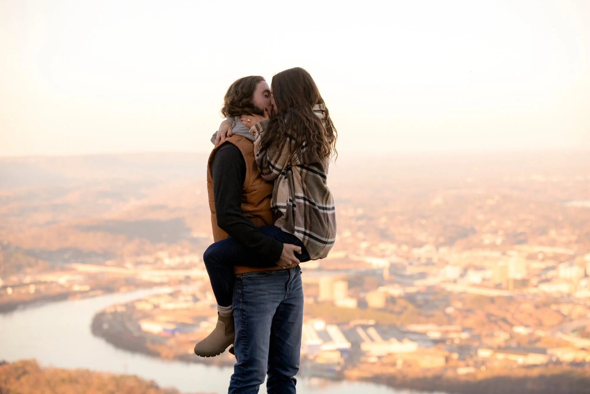 A couple kissing, with the woman sitting on the man's arms, overlooking a cityscape at sunset.