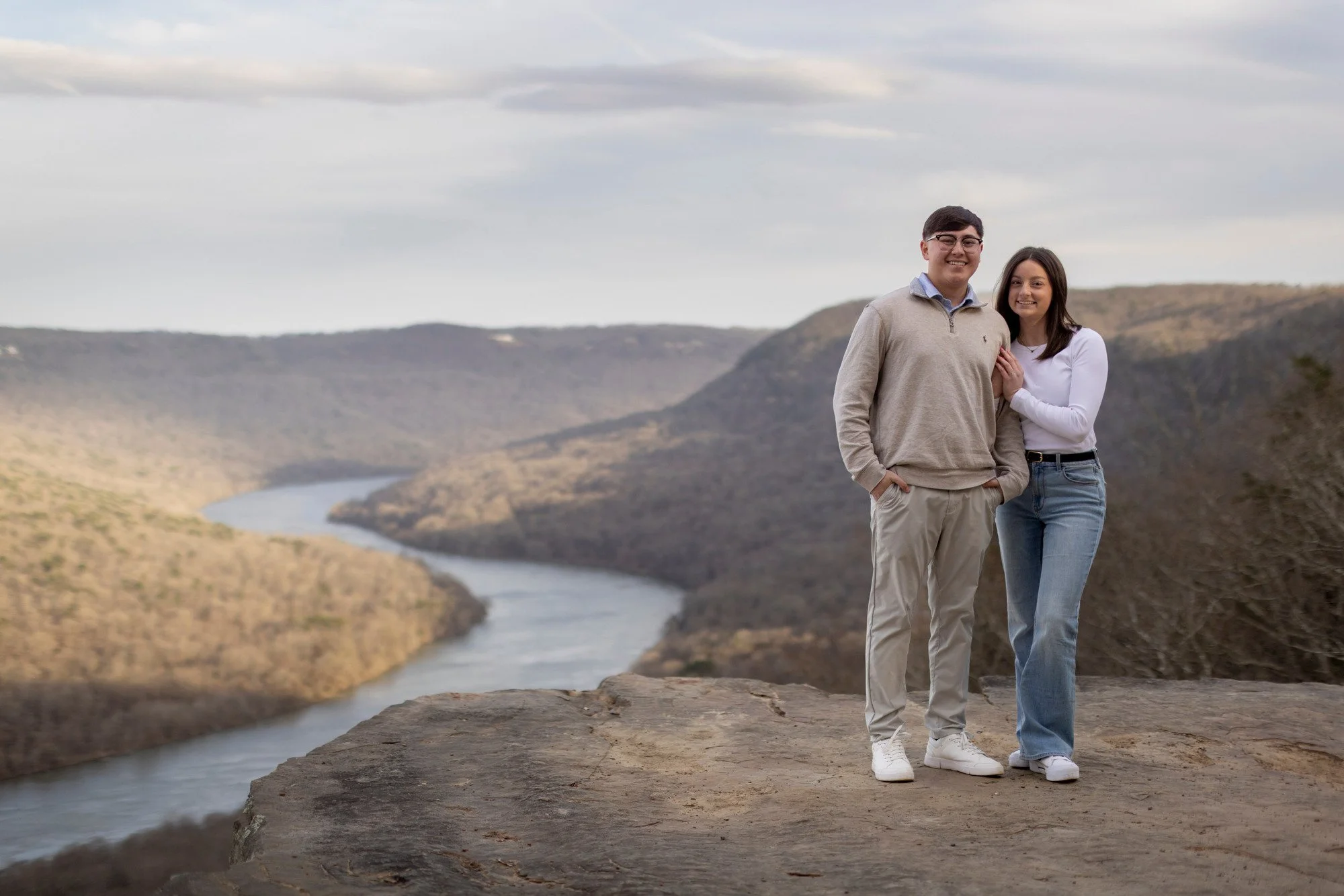 A smiling young man and woman standing together outdoors near a river with hills in the background. The man is wearing glasses, a beige sweater, and beige pants. The woman is wearing a white long-sleeve shirt and blue jeans. They are holding hands an