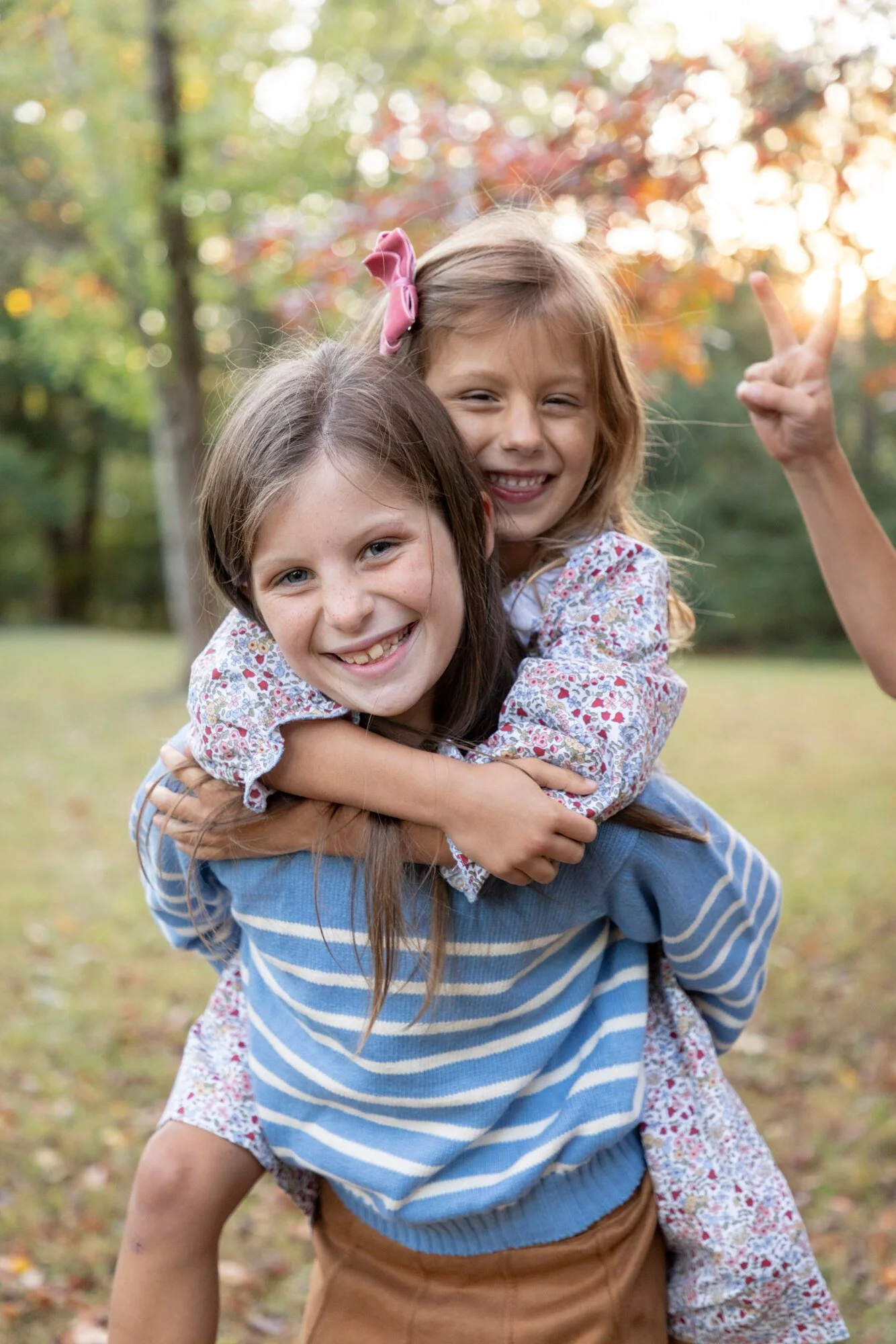 Two young girls smiling and hugging each other outdoors during fall, with trees and colorful leaves in the background.