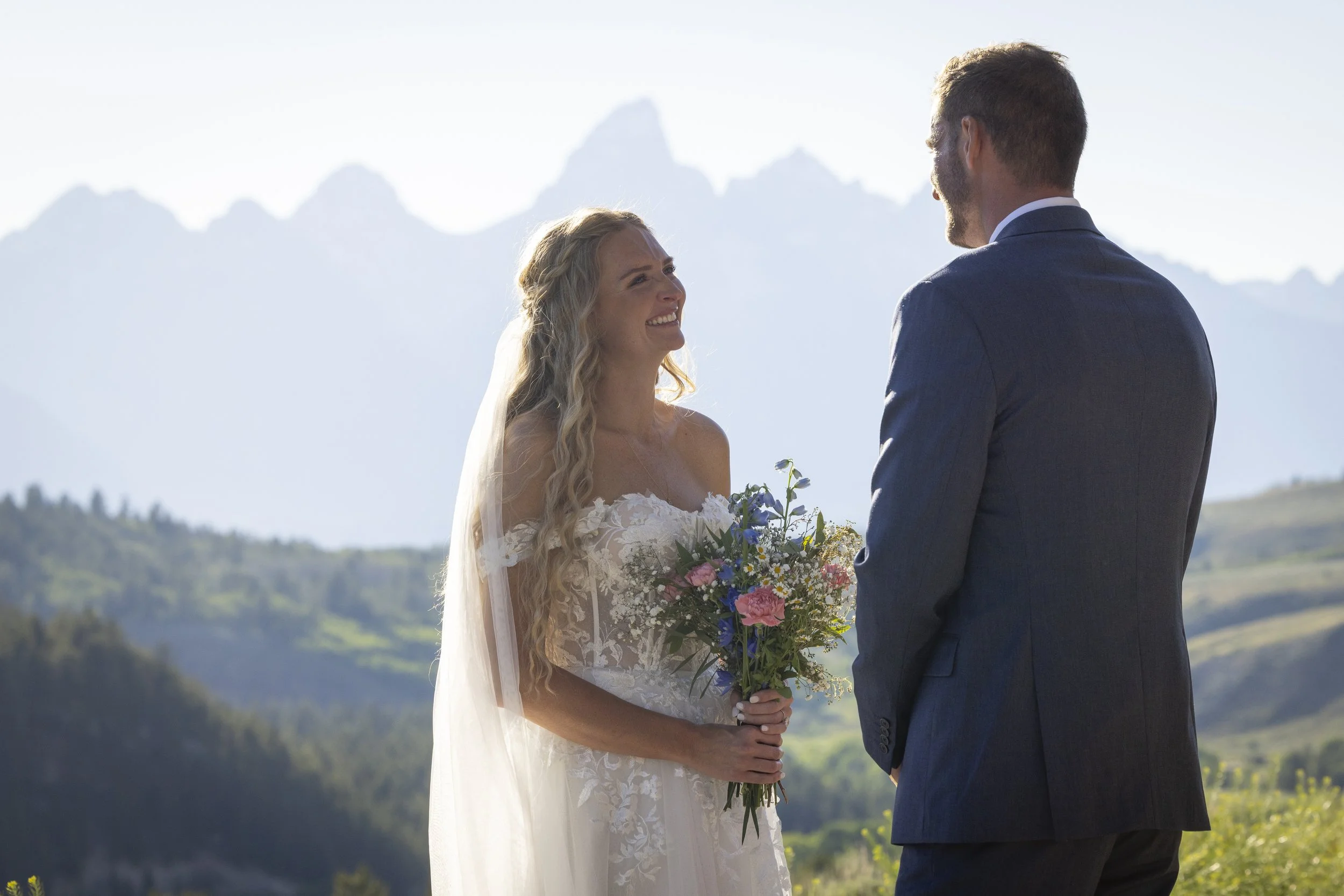 A bride and groom standing outdoors during their wedding ceremony with mountains in the background. The bride is wearing a white lace wedding dress and holding a bouquet of flowers, smiling at the groom, who is wearing a dark suit.