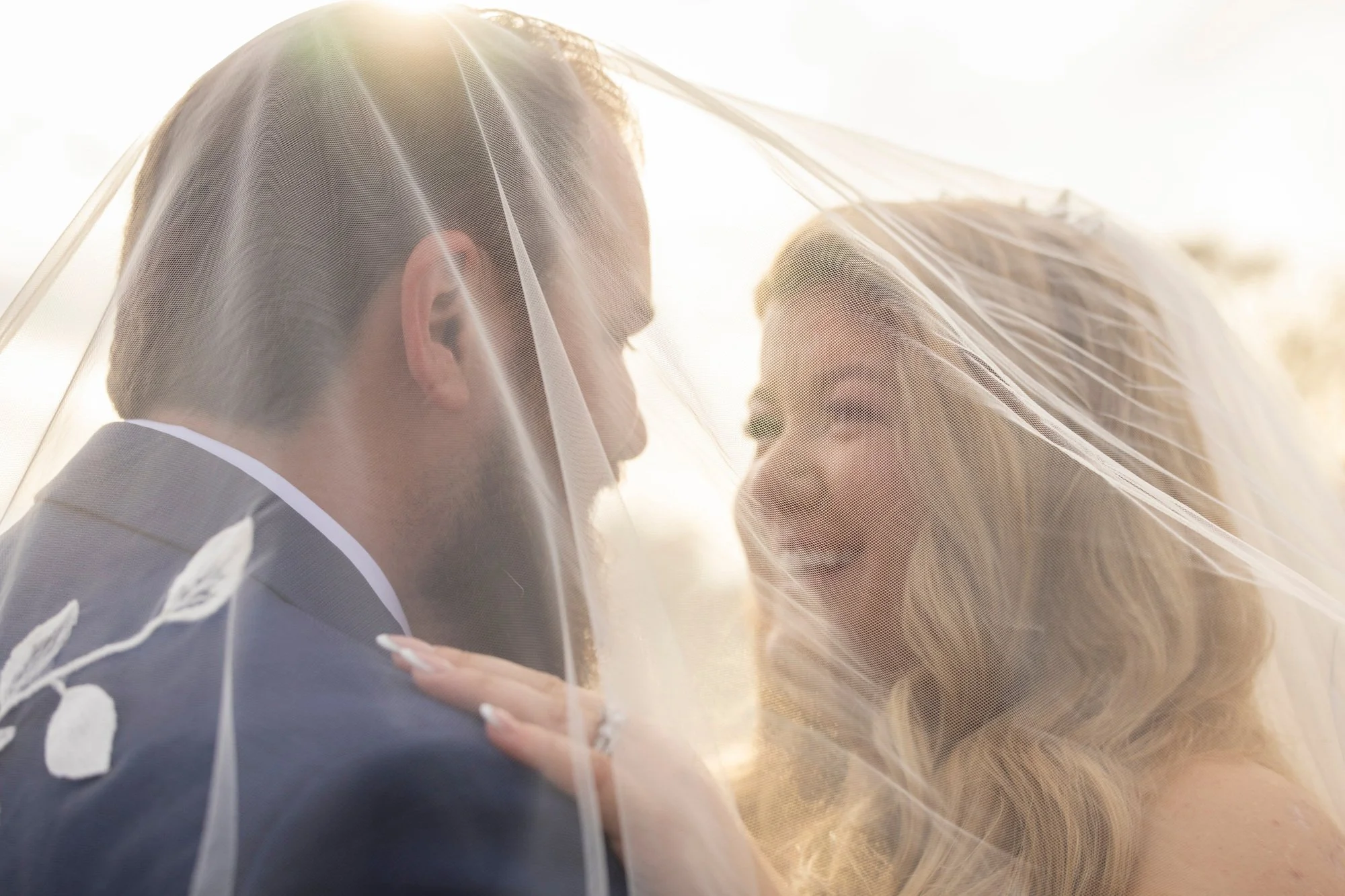 A close-up of a bride and groom with a veil covering their faces, sharing an intimate moment outdoors with warm sunlight.