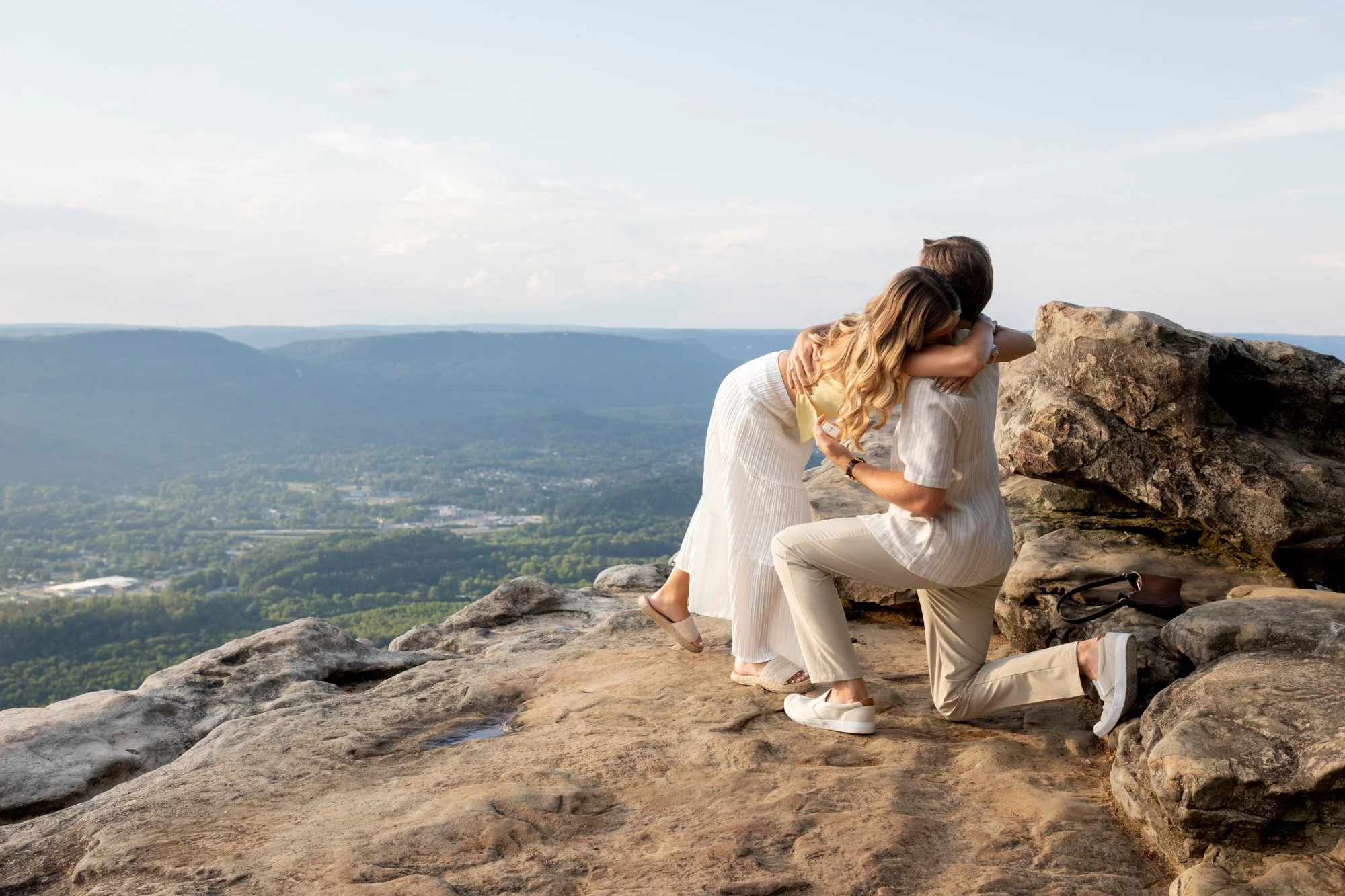 A man kneeling on a rock embracing and proposing marriage to a woman on a mountain overlook with a scenic view of the valley and distant mountains.