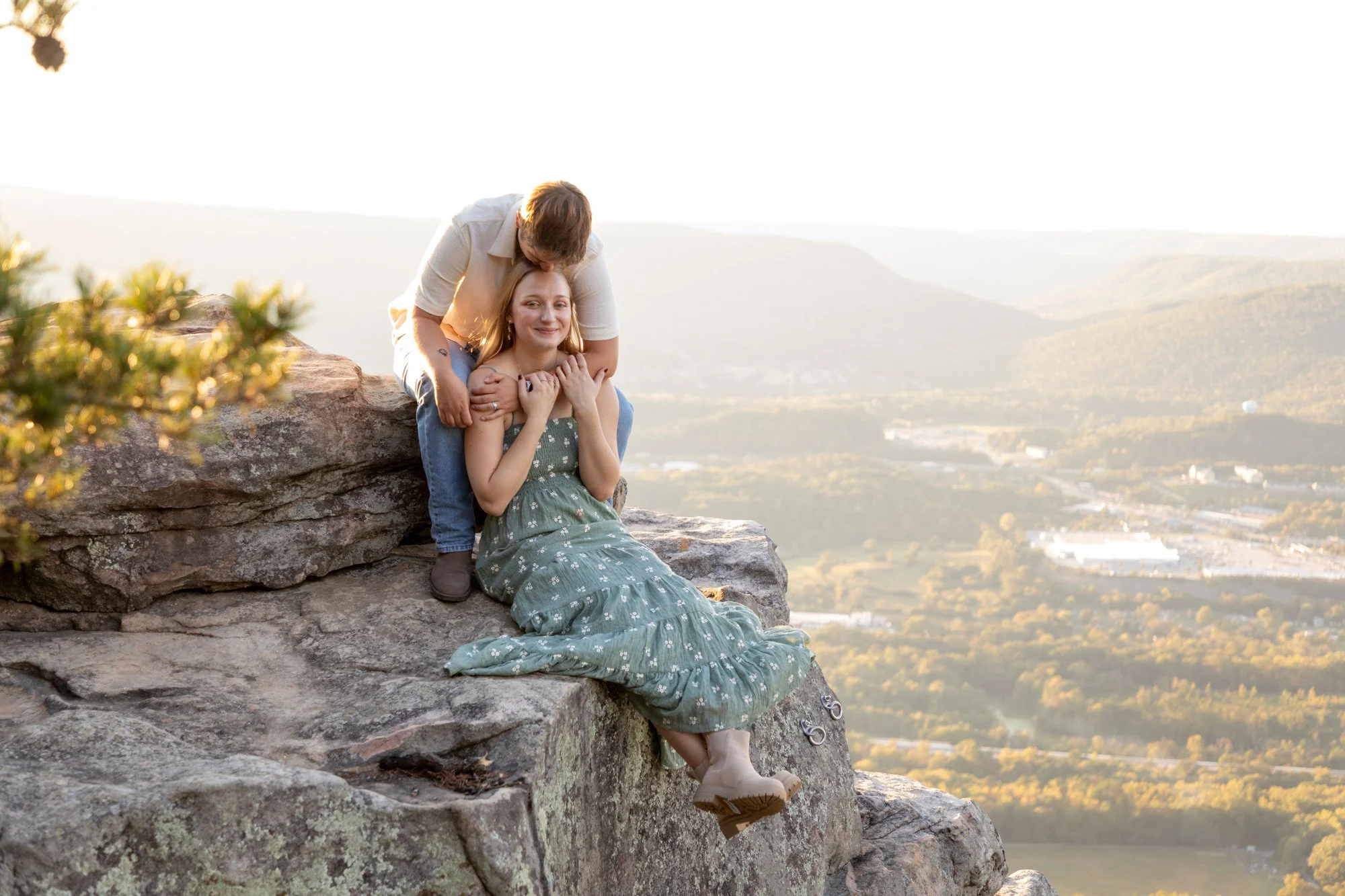 A couple sitting on a rocky ledge overlooking a scenic valley at sunset. The woman is smiling and sitting with her legs crossed, wearing a green floral dress and beige boots. The man is leaning over her, kissing her on the forehead, with his arms aro