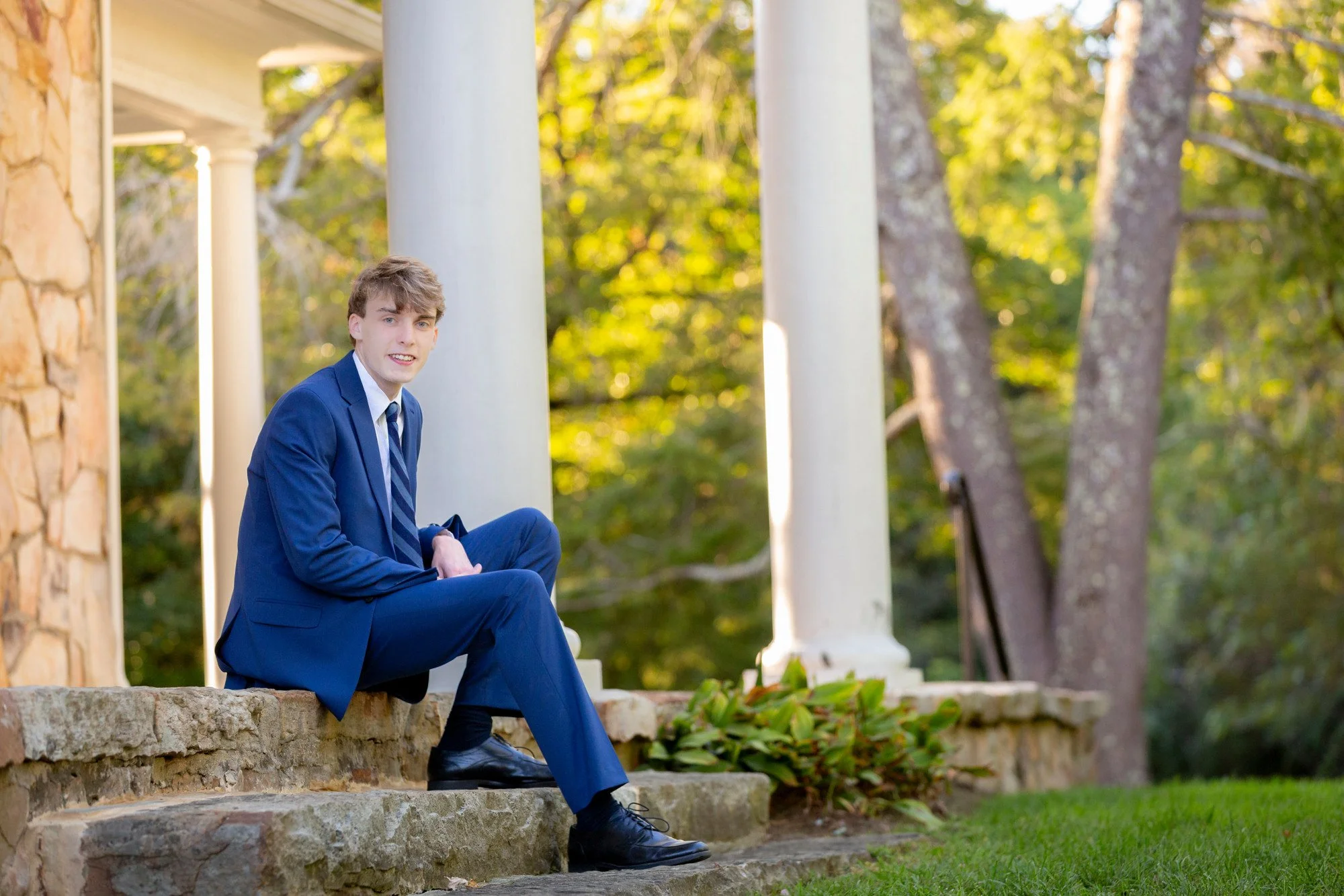 A young man in a blue suit sitting on stone steps outdoors surrounded by trees and greenery.