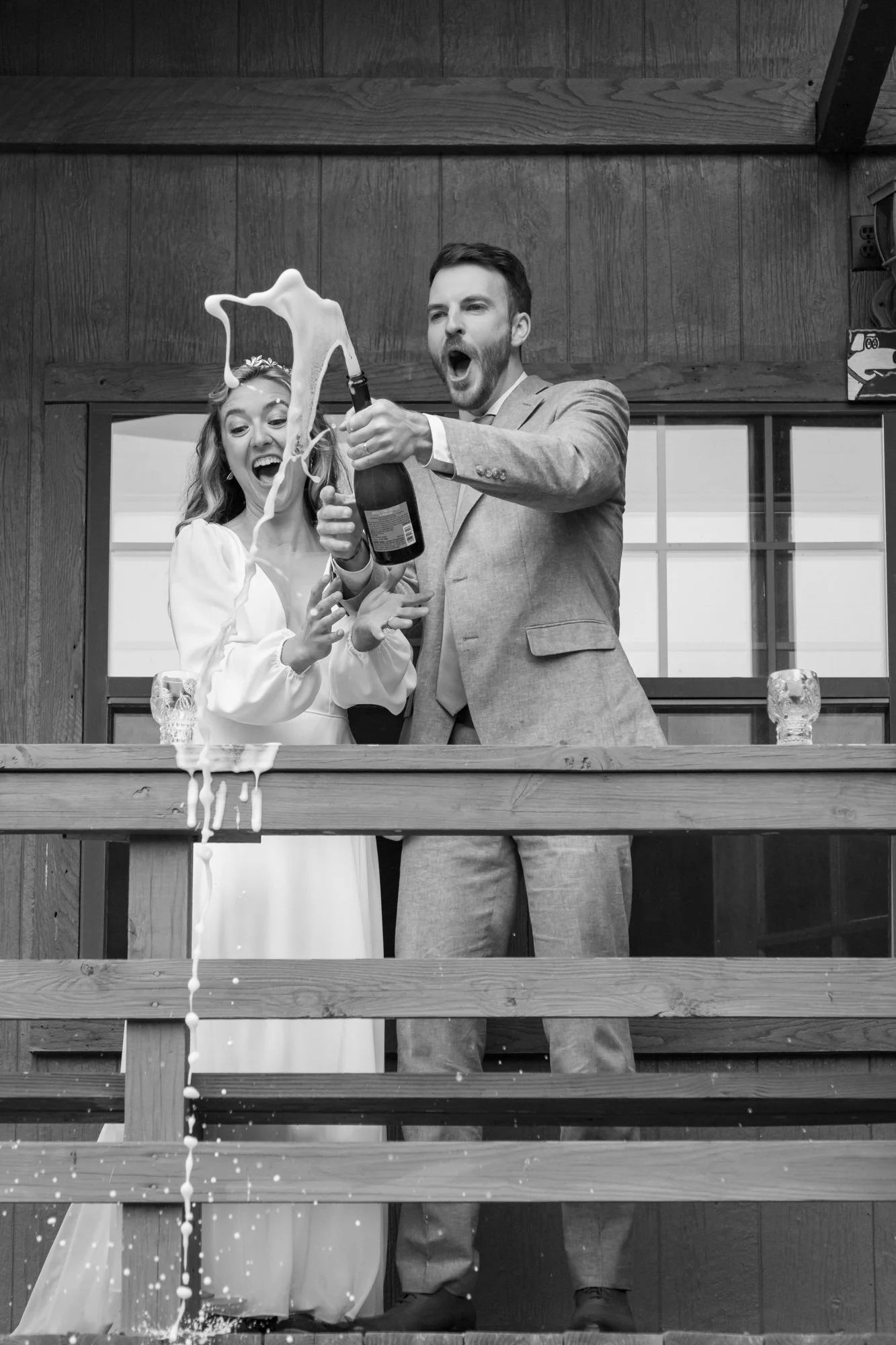 A black and white photograph of a wedding reception scene with a bride and groom standing on a wooden balcony. The bride is dressed in a white gown with long sleeves and a tiara, and the groom is in a gray suit with a light shirt. They are both smili