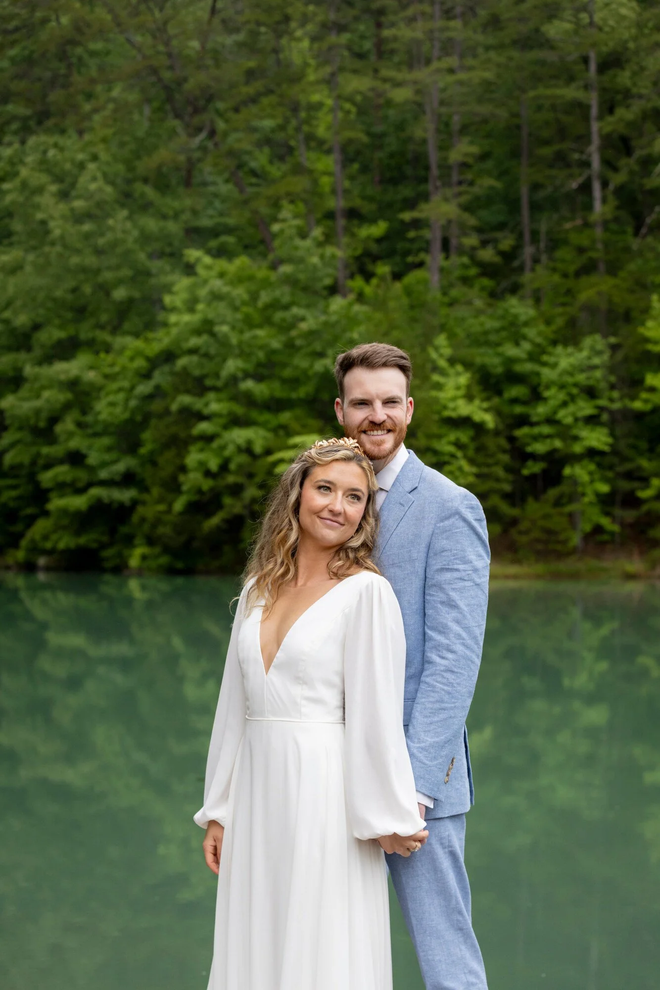 A couple dressed in formal wedding attire standing outdoors near a body of water with a forest background.