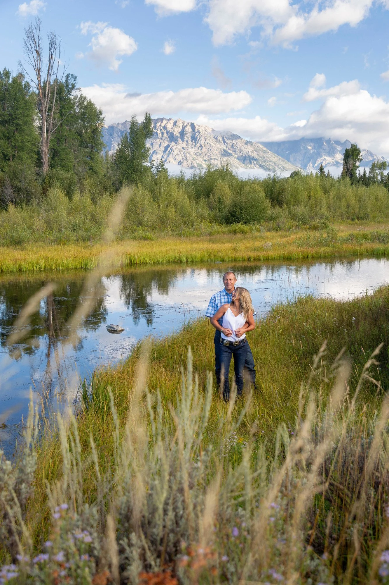 A couple standing by a river in a scenic natural landscape with trees, mountains, and a blue sky with clouds.