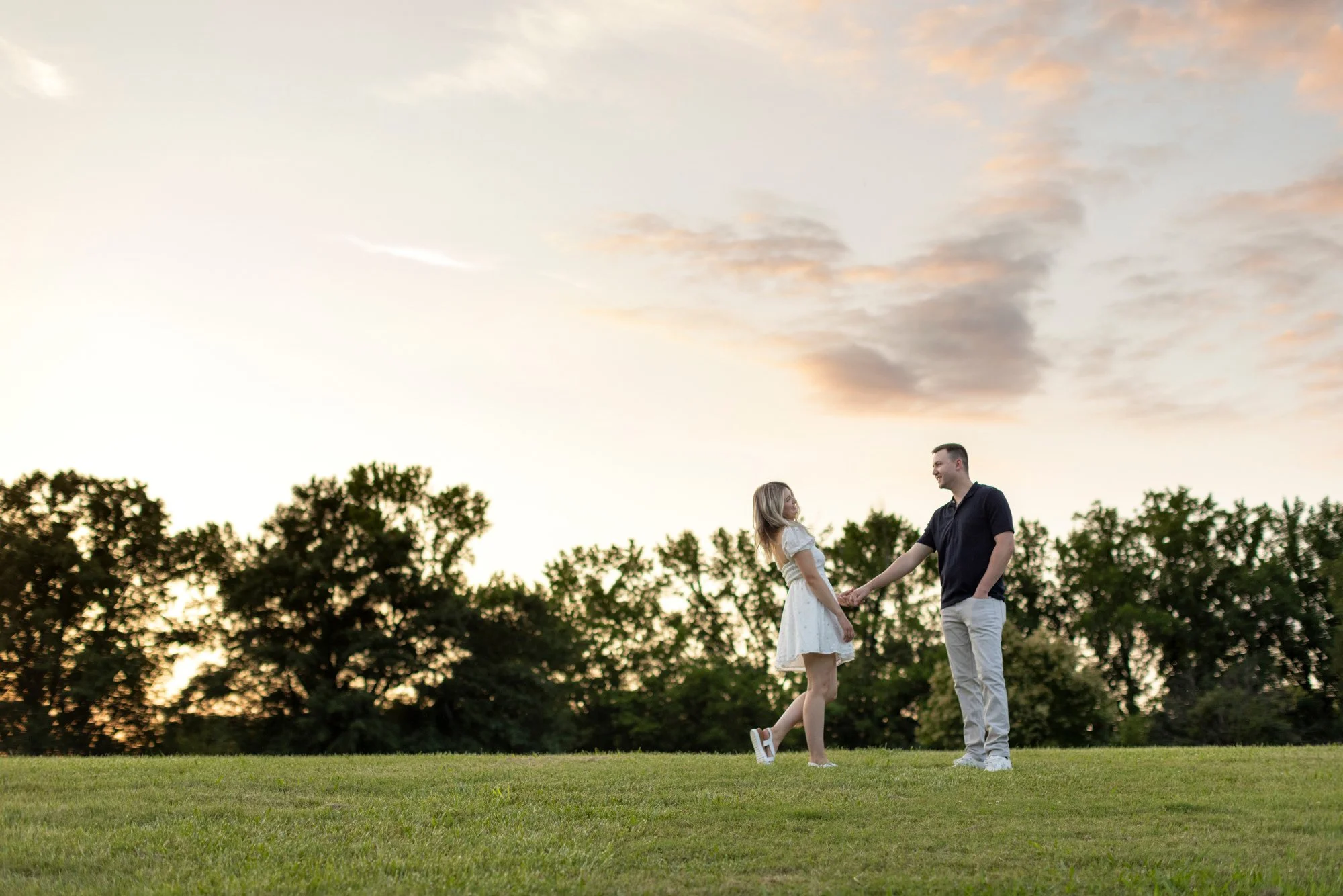 A young couple holding hands and enjoying a moment outdoors during sunset, with the sky partly cloudy and trees in the background.