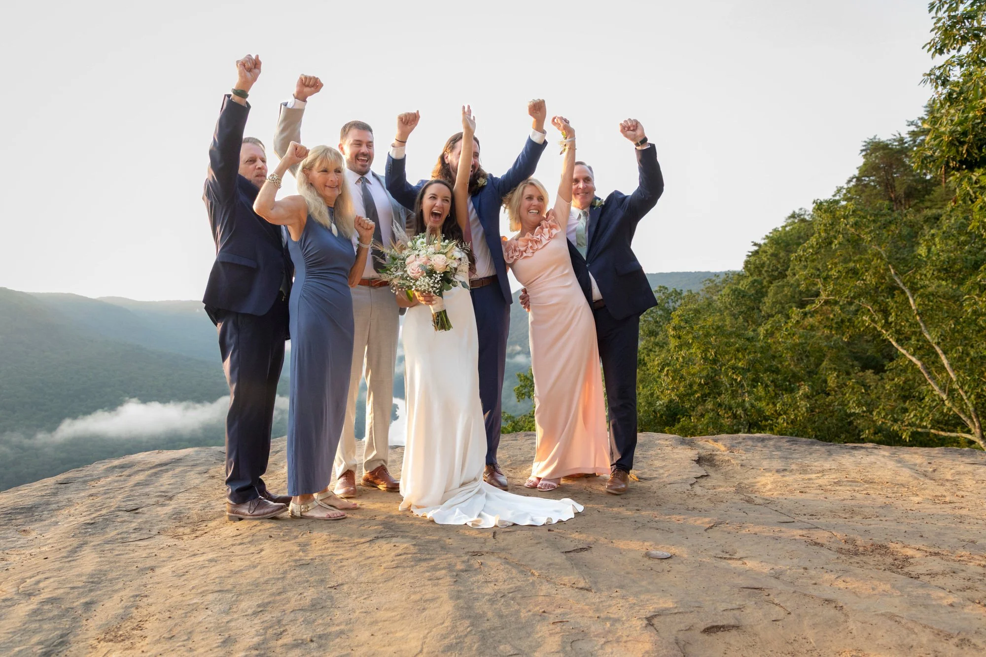 Group of seven people celebrating on a rocky outdoor ledge, with a scenic mountain landscape and clear sky in the background. The group includes a bride in a white gown holding a bouquet, surrounded by guests in formal attire, all with raised fists a