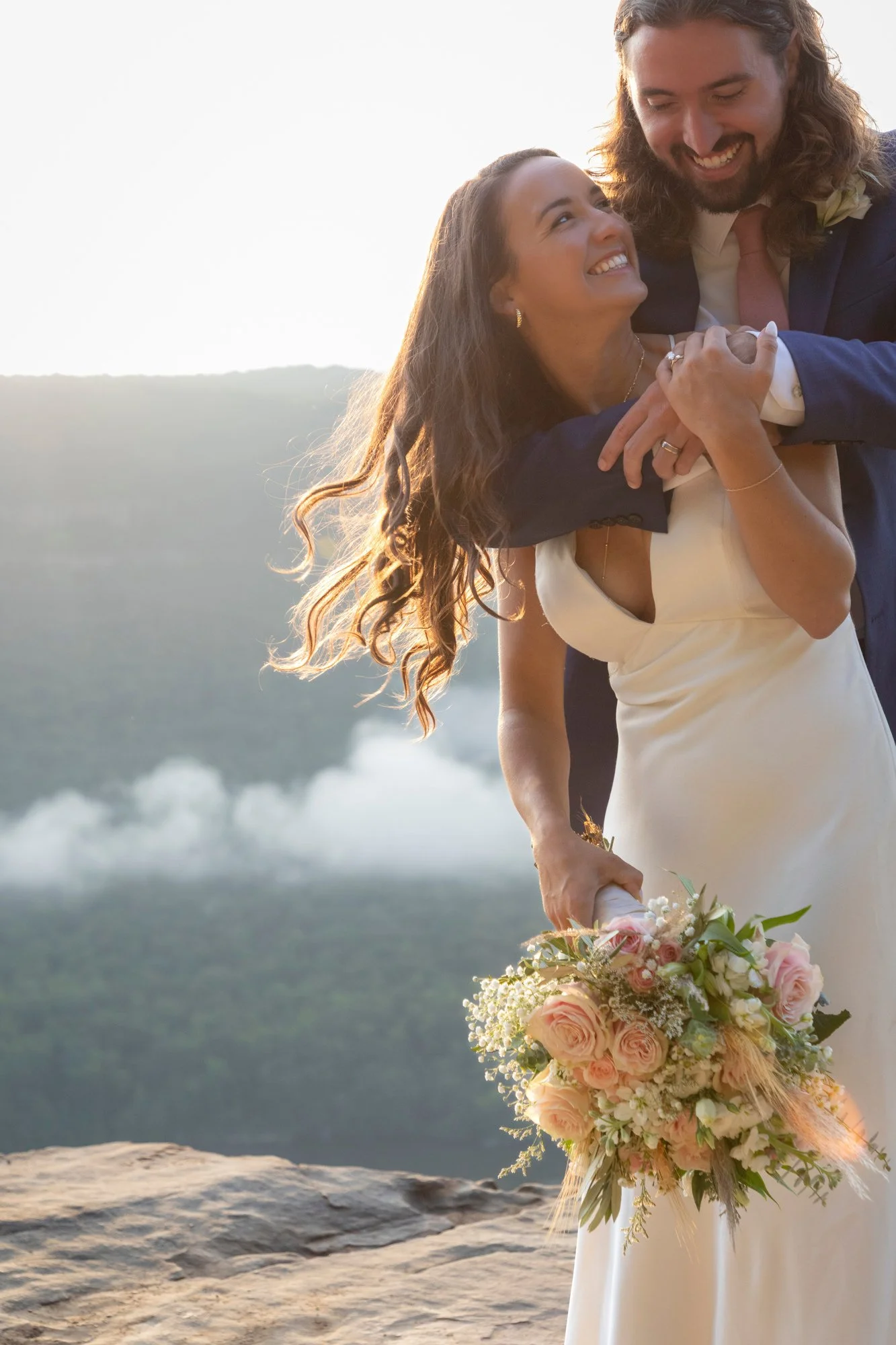 A bride and groom embrace outdoors in front of a scenic landscape with water and mountains, during sunset. The bride holds a bouquet of pink and white flowers.