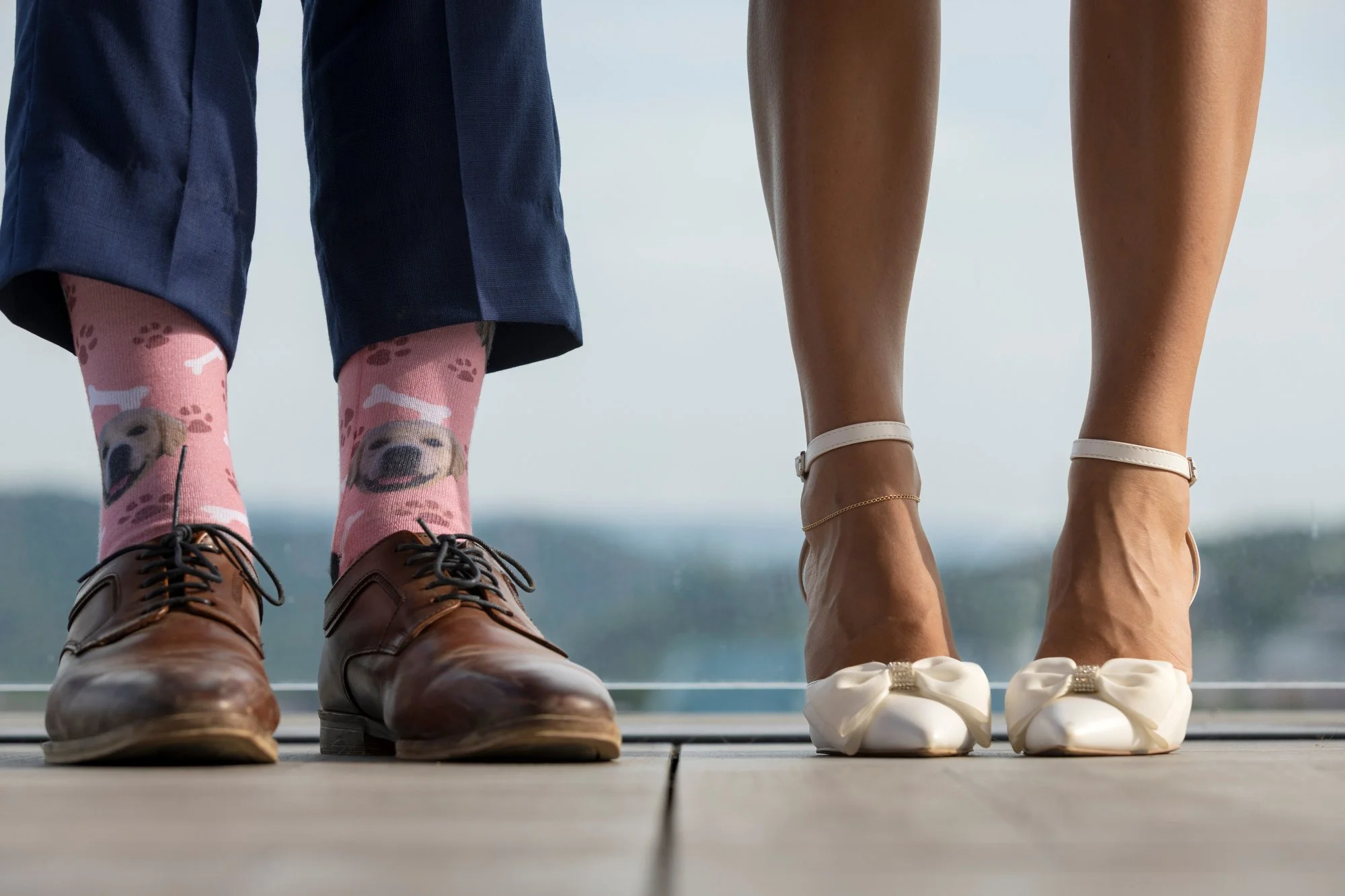 Close-up of the feet and legs of two women standing side by side outdoors on a wooden deck. The woman on the left wears brown leather shoes, pink socks with a dog pattern, and dark blue pants. The woman on the right wears white sneakers, a delicate a