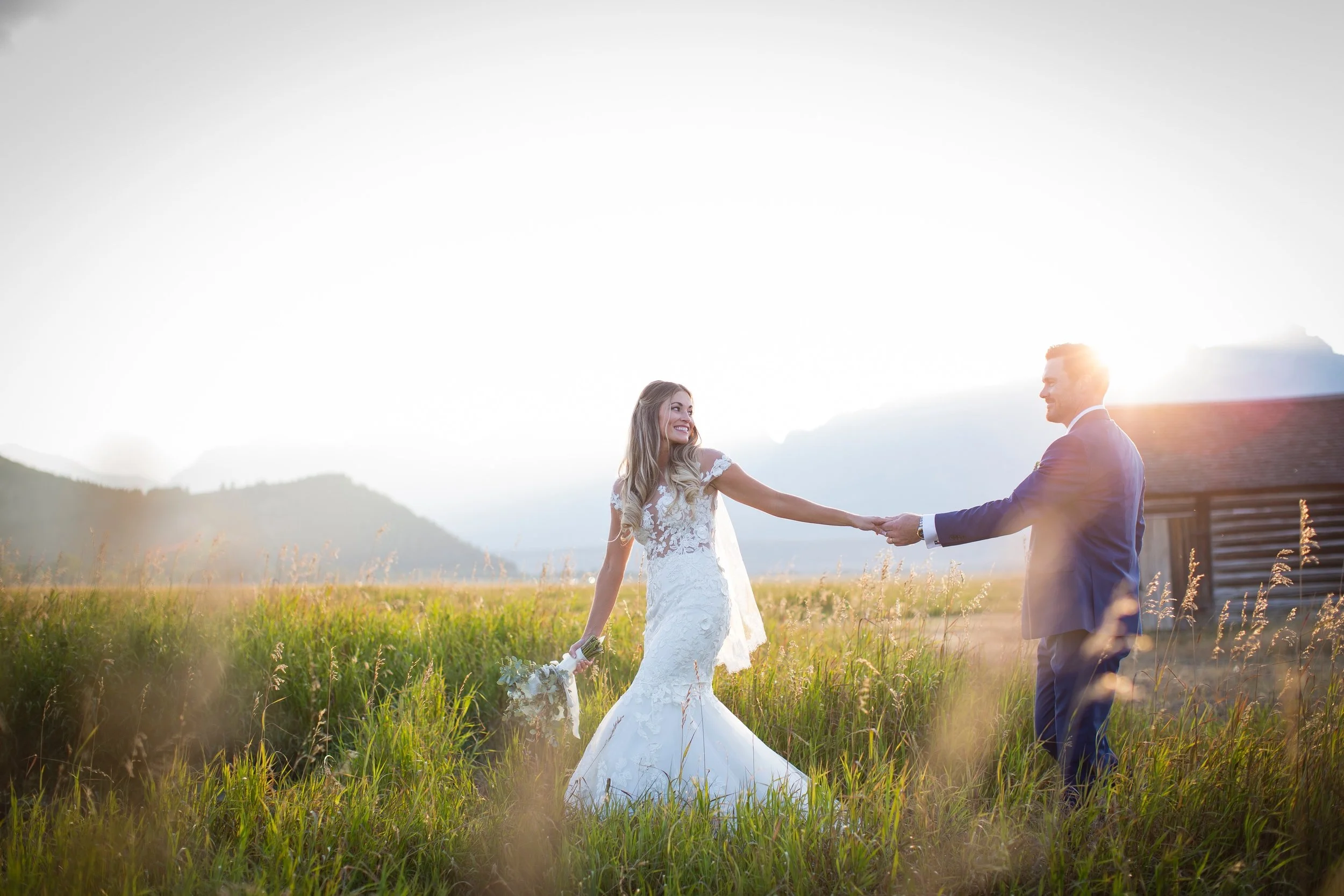 A bride and groom holding hands in a field at sunset, with mountains in the background.