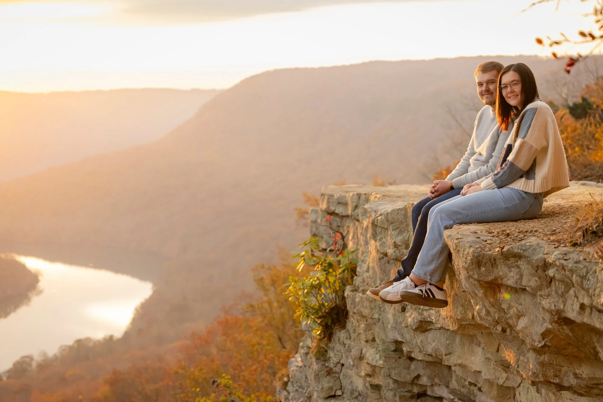 A man and a woman sit on a stone ledge overlooking a river and mountains during sunset. They are smiling and wearing casual sweaters and jeans.
