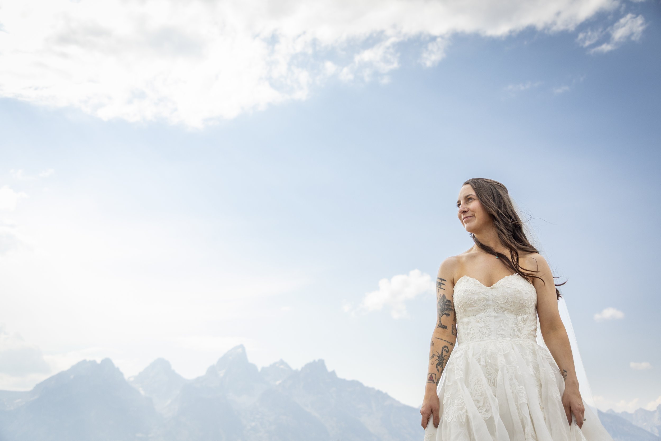 A woman with long brown hair, tattoos on her arms, and wearing a white lace dress standing outdoors against a backdrop of mountains and a bright blue sky with scattered clouds.
