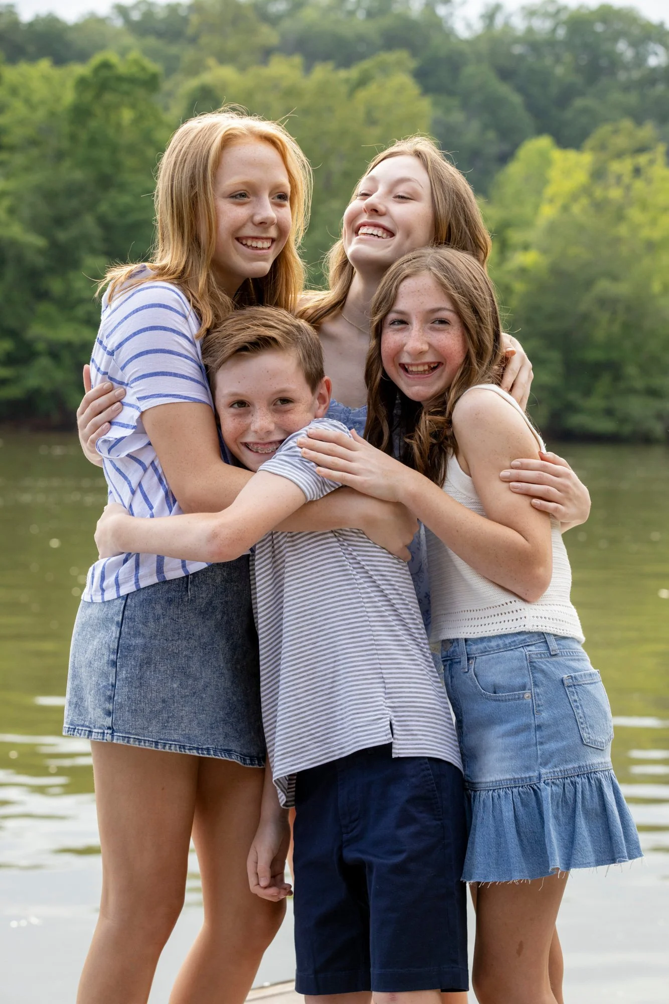 A group of five children, three girls and two boys, hugging each other outdoors by a lake with a green, forested background. They are smiling and enjoying a day outside.