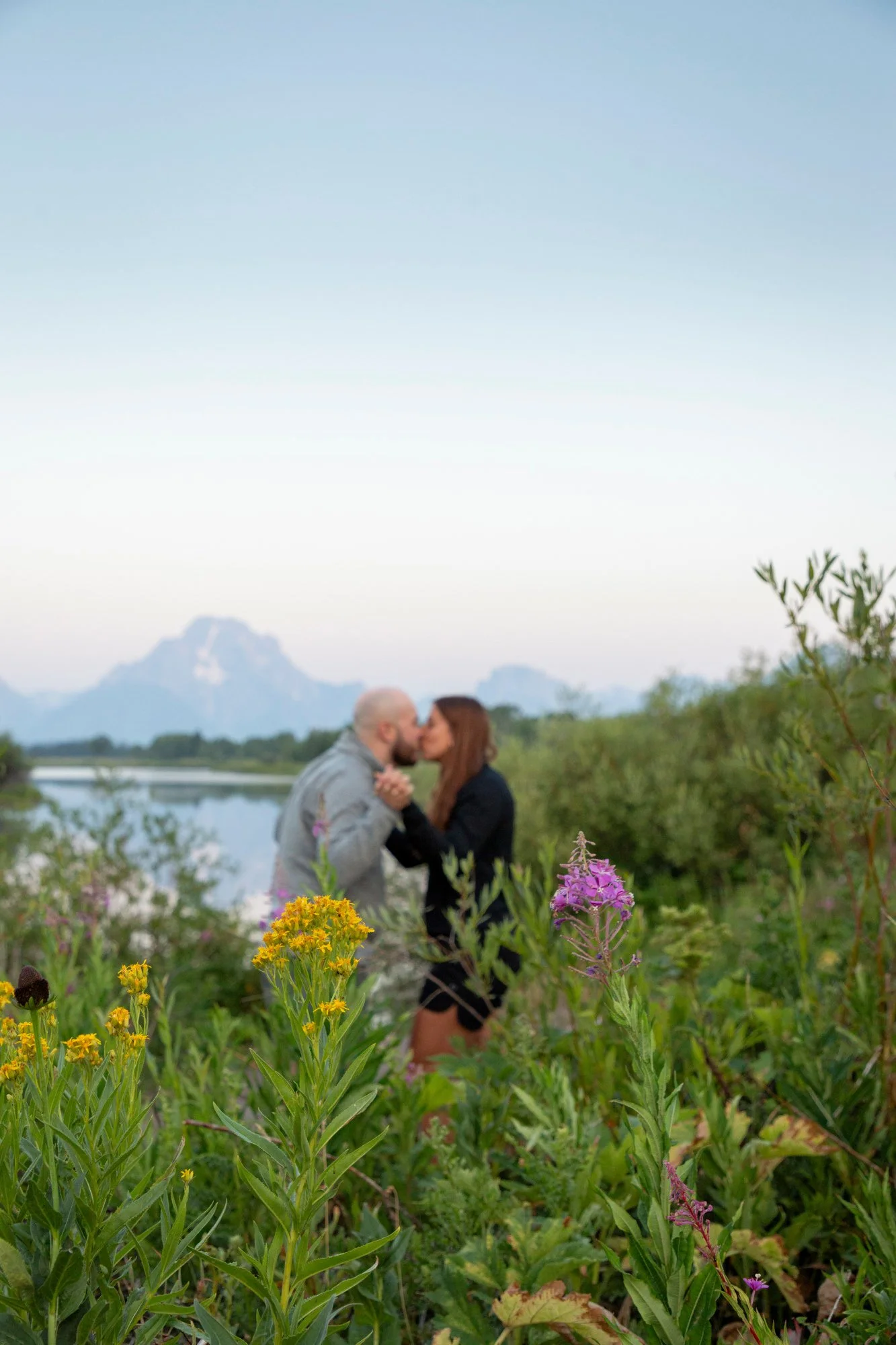 A couple sharing a kiss in a lush green outdoor setting with mountains in the background, surrounded by colorful wildflowers in the foreground.