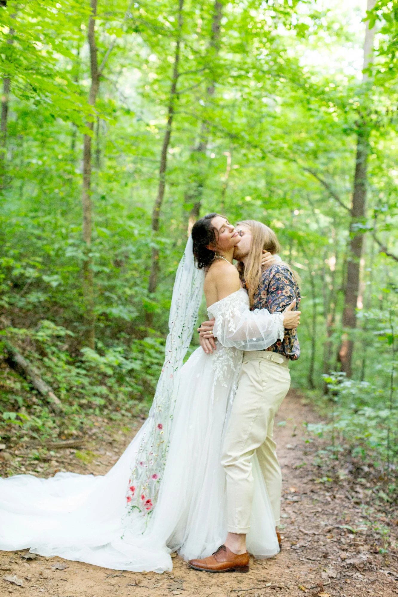 Gay couple kissing in a forest, one wearing a wedding dress and the other wearing cream-colored pants and a floral shirt.