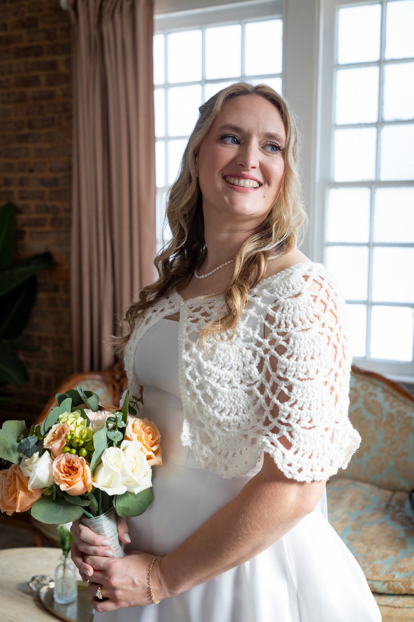 A bride in a white dress with a crochet jacket holding a bouquet of orange and white roses, in a room with large windows and brick walls.