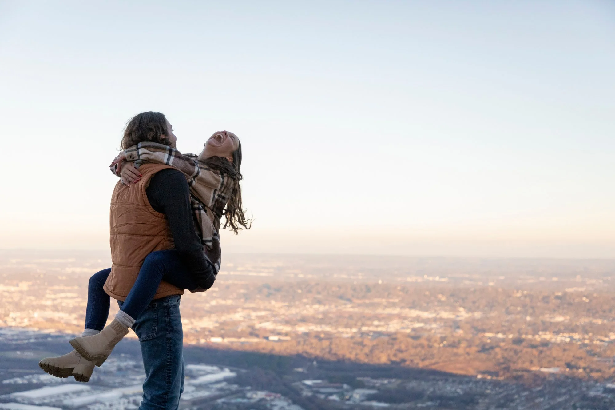 A man carrying a woman on his back while standing on a mountain or hilltop during sunrise or sunset, with a view of a city below.
