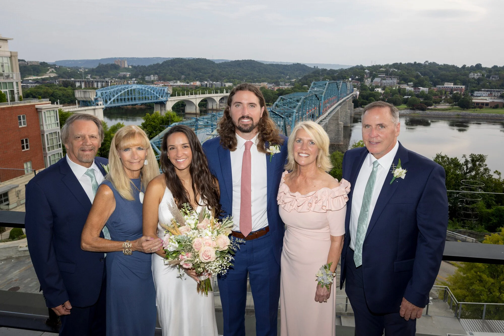 A group of seven people dressed in formal attire, standing on an outdoor balcony with a scenic river and bridge in the background. One woman is holding a bouquet of flowers, indicating a celebration or special occasion.