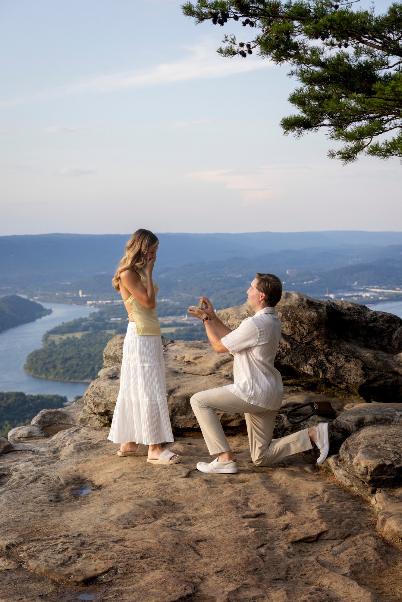 A man proposing marriage to a woman on a scenic rocky ledge overlooking a river and mountains, with both dressed in light-colored clothing and surrounded by nature.