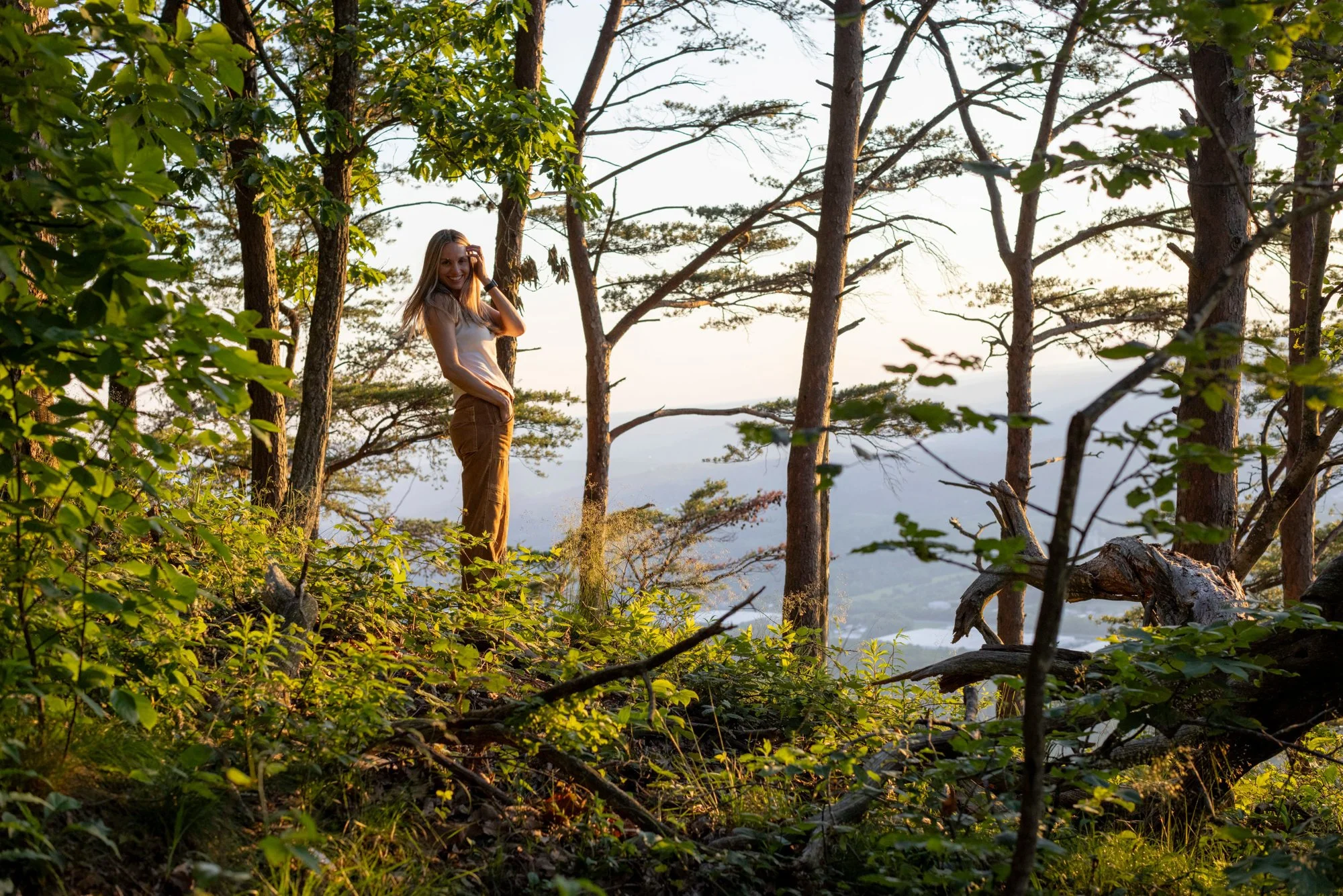 A woman with long blonde hair stands among trees and greenery during sunset, smiling and touching her hair.