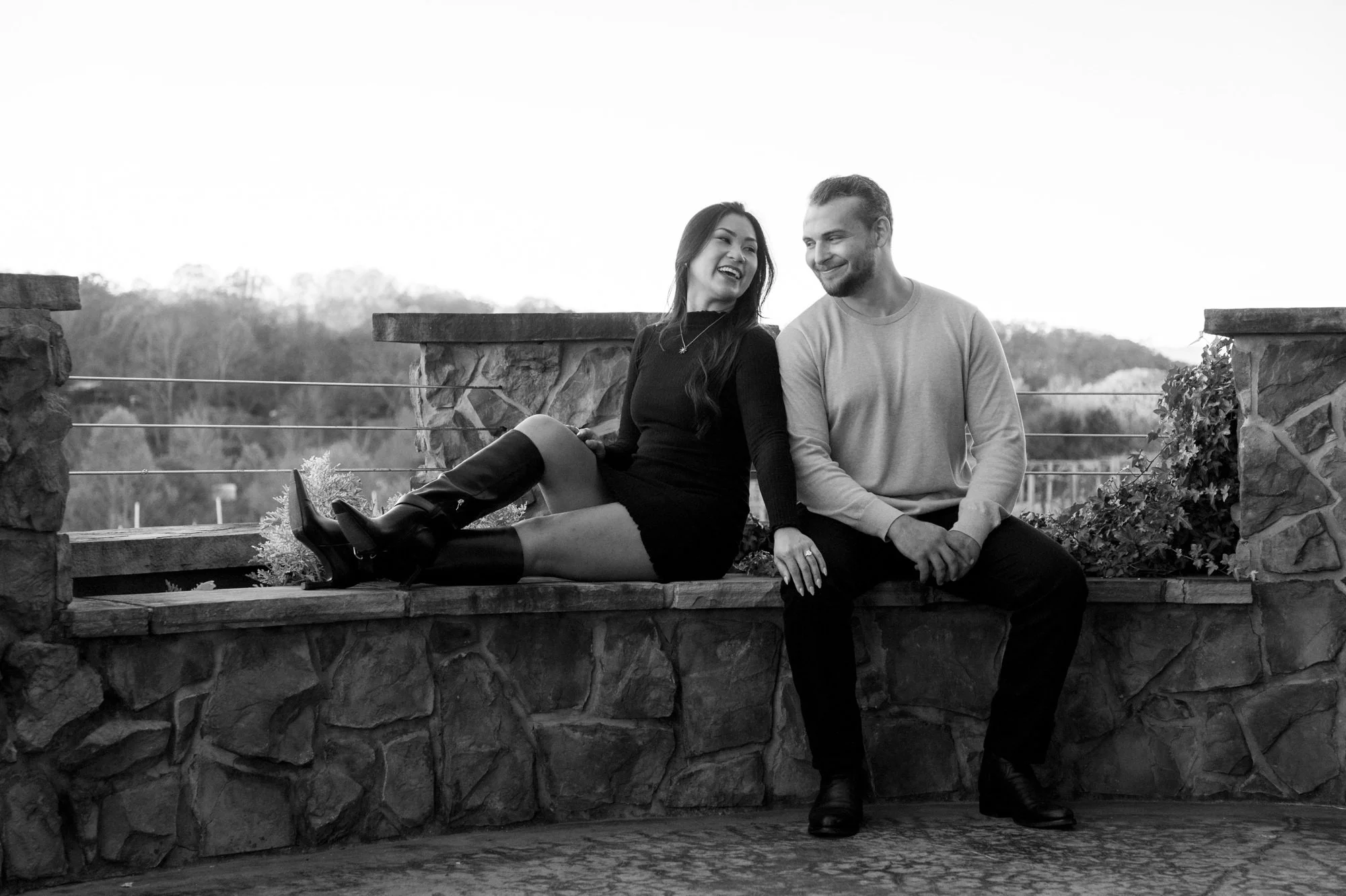 A black and white photo of a happy couple sitting and leaning on a stone railing outdoors with trees in the background, smiling at each other.
