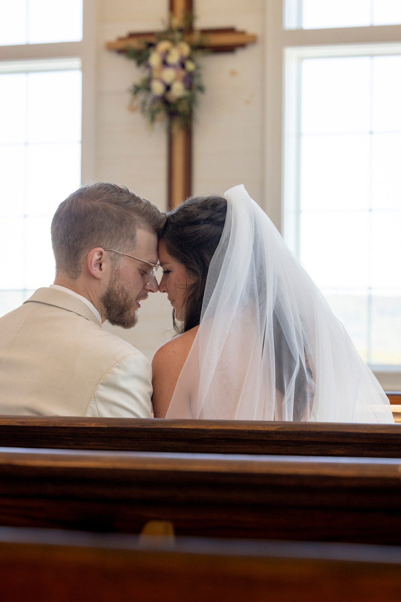A couple at a wedding in a church, with heads touching, in front of a cross decorated with flowers, and large windows in the background.