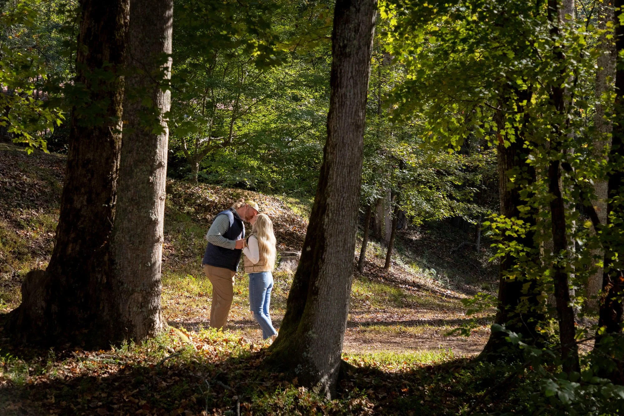 A man and a young girl stand close together in a forest with tall trees and lush green foliage, sunlight filtering through the leaves, as they appear to be sharing a moment.