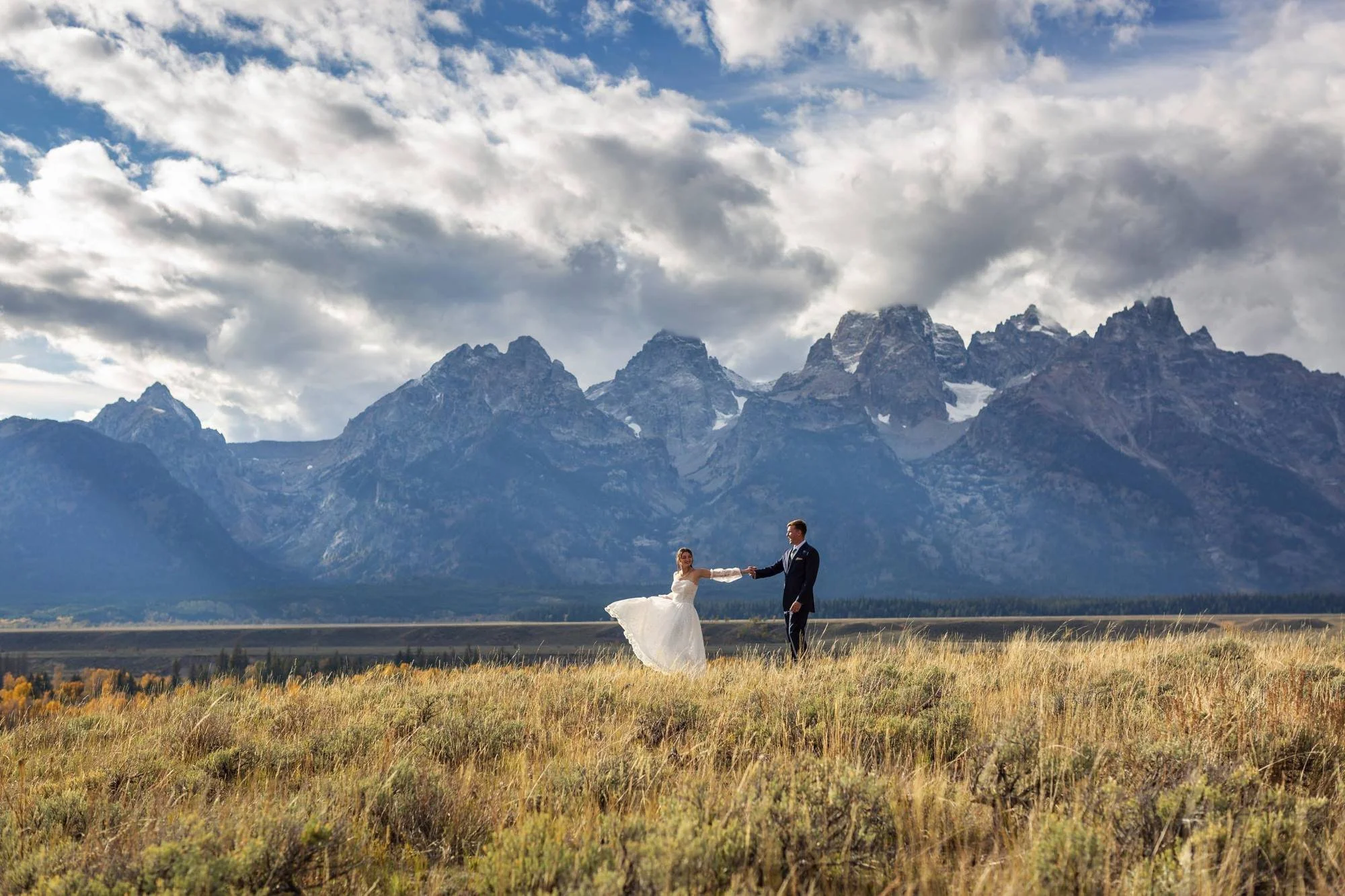 A bride and groom holding hands in a field with mountains and clouds in the background.