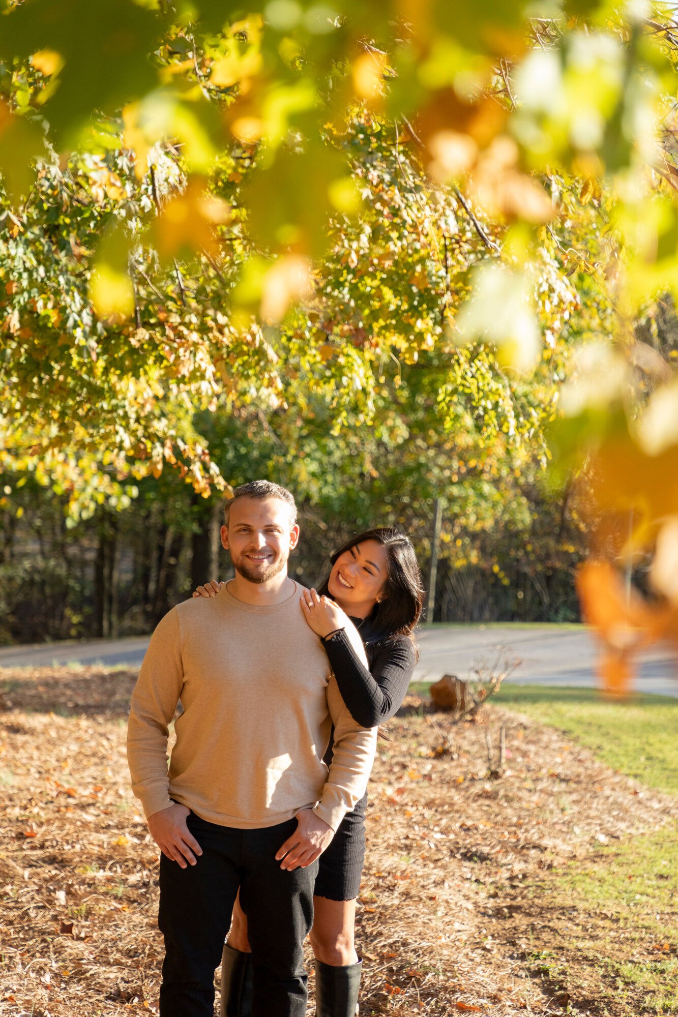 A man and a woman smiling and standing close together outdoors on a sunny day with colorful autumn leaves around.