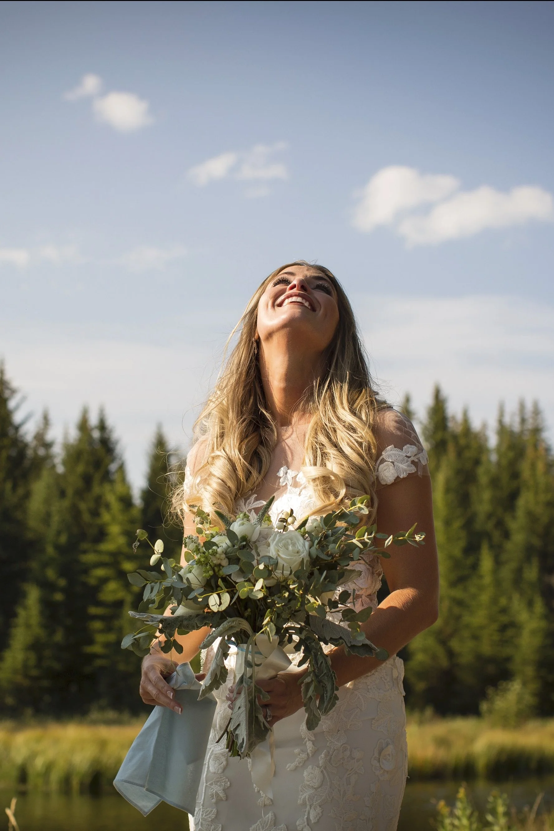 A smiling woman in a white lace dress holding a bouquet of white roses and greenery outdoors against a backdrop of trees and blue sky with clouds.
