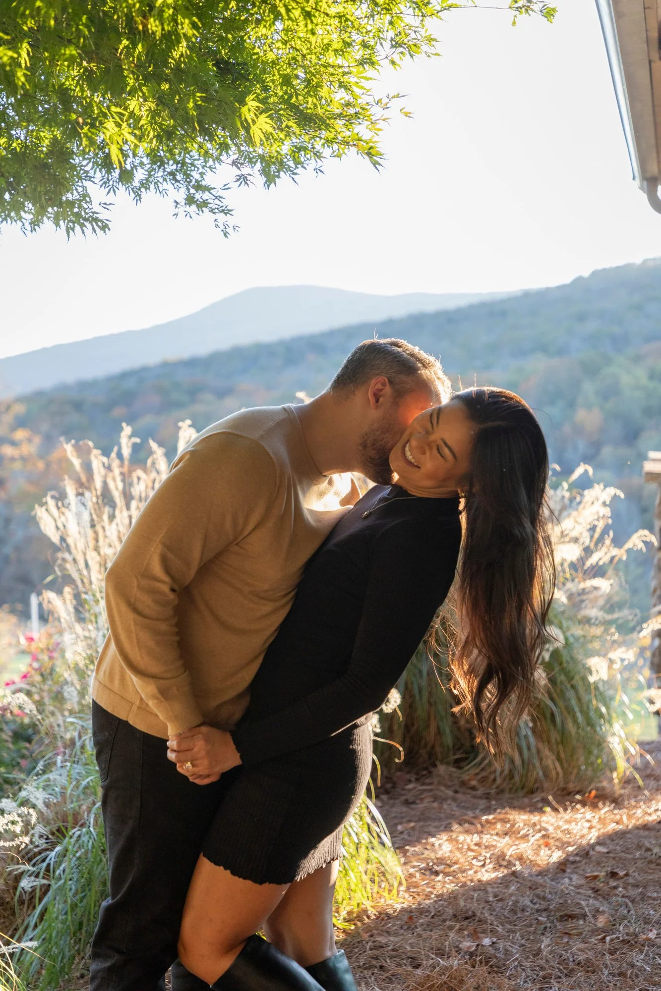 A couple standing outdoors, smiling and sharing a kiss, with trees, mountains, and bright sunlight in the background.