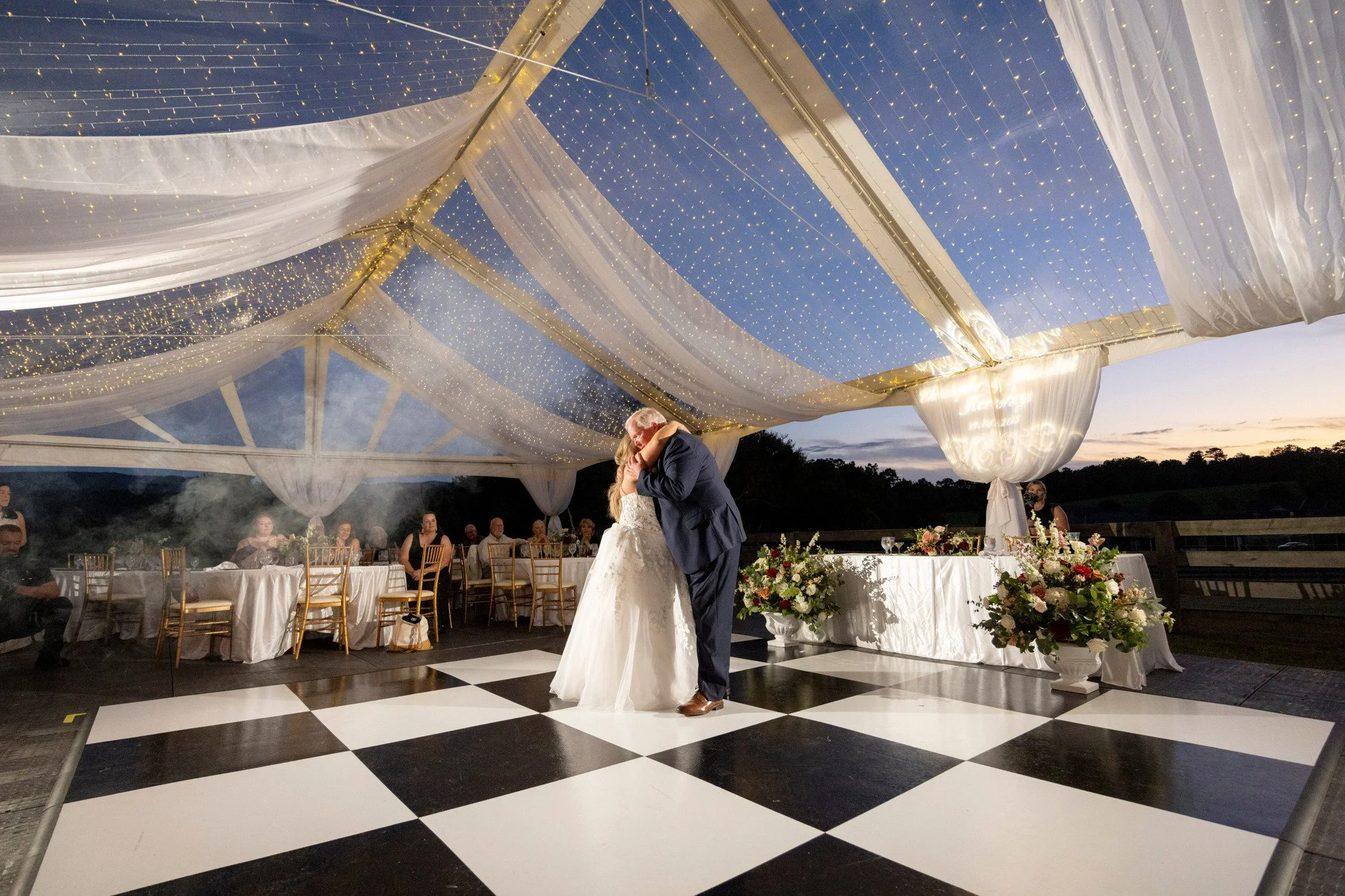 A bride and groom dancing together under a tent at sunset with draped white fabric and string lights, with wedding guests seated at decorated tables nearby.