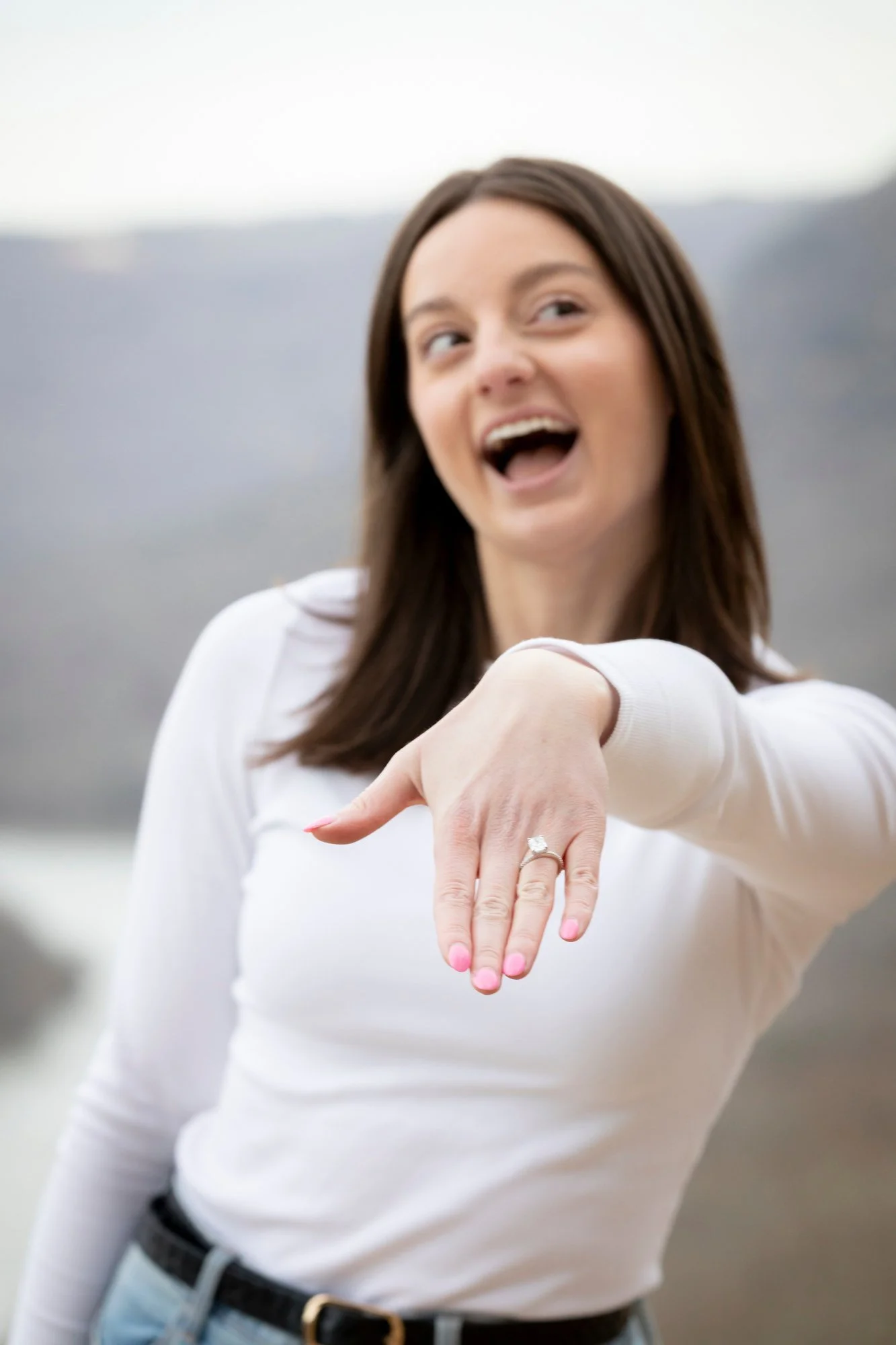 A woman smiling and showing a wedding ring on her finger with her arm extended, outdoors with a blurry landscape background.